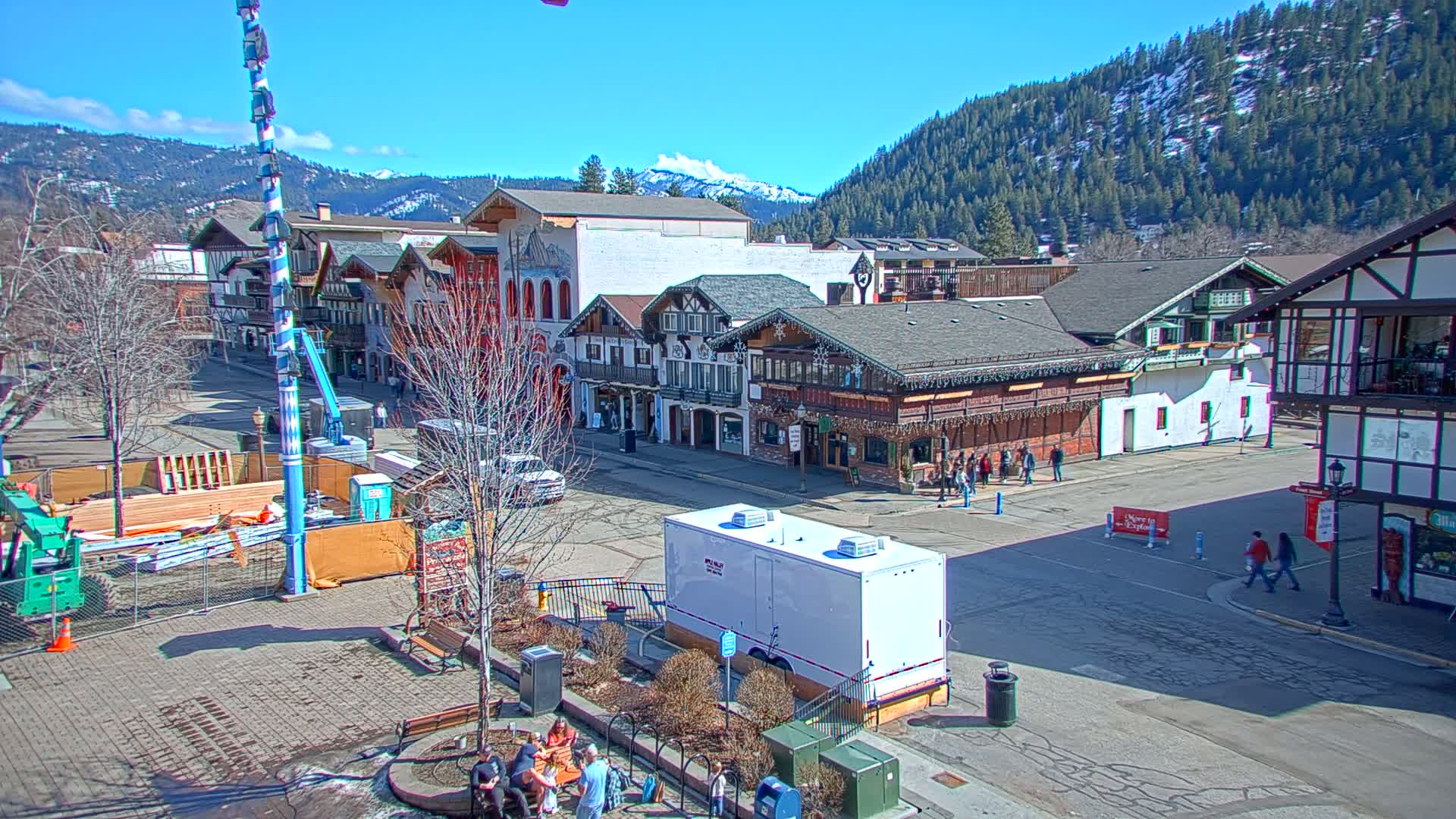 A town square with several buildings in a European architectural style sits against a backdrop of snow-capped mountains under a clear blue sky.