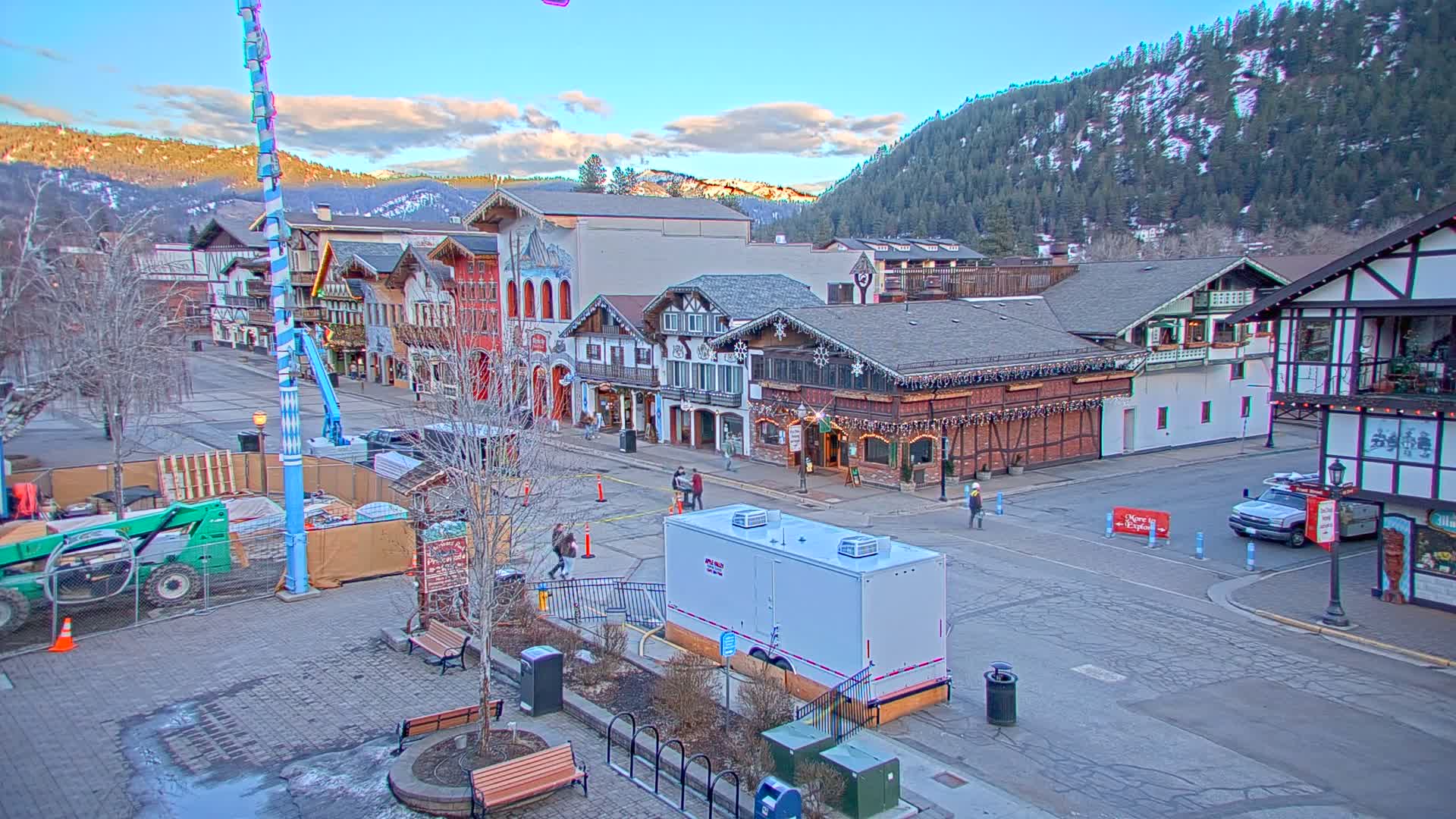 A mostly empty town square with several European-style buildings, construction equipment, and a few pedestrians, under a partly cloudy sky.