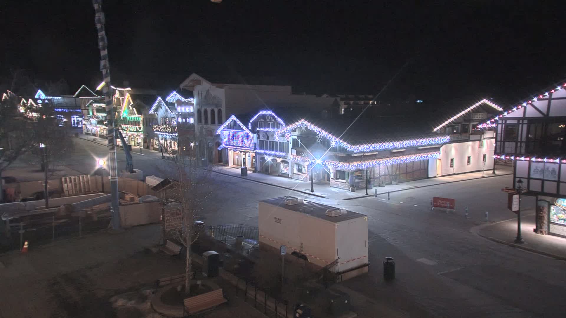 A nighttime, snow-covered street scene shows a row of buildings decorated with strings of lights.