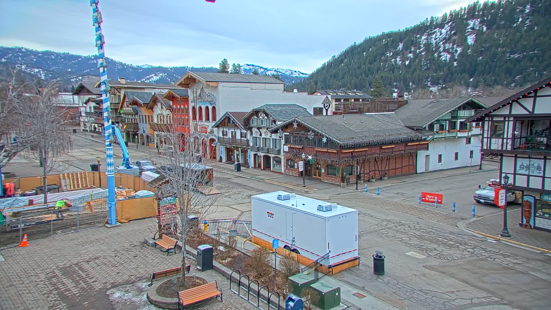 A town square with Bavarian-style buildings is under construction, with overcast skies and snow-covered mountains in the background.
