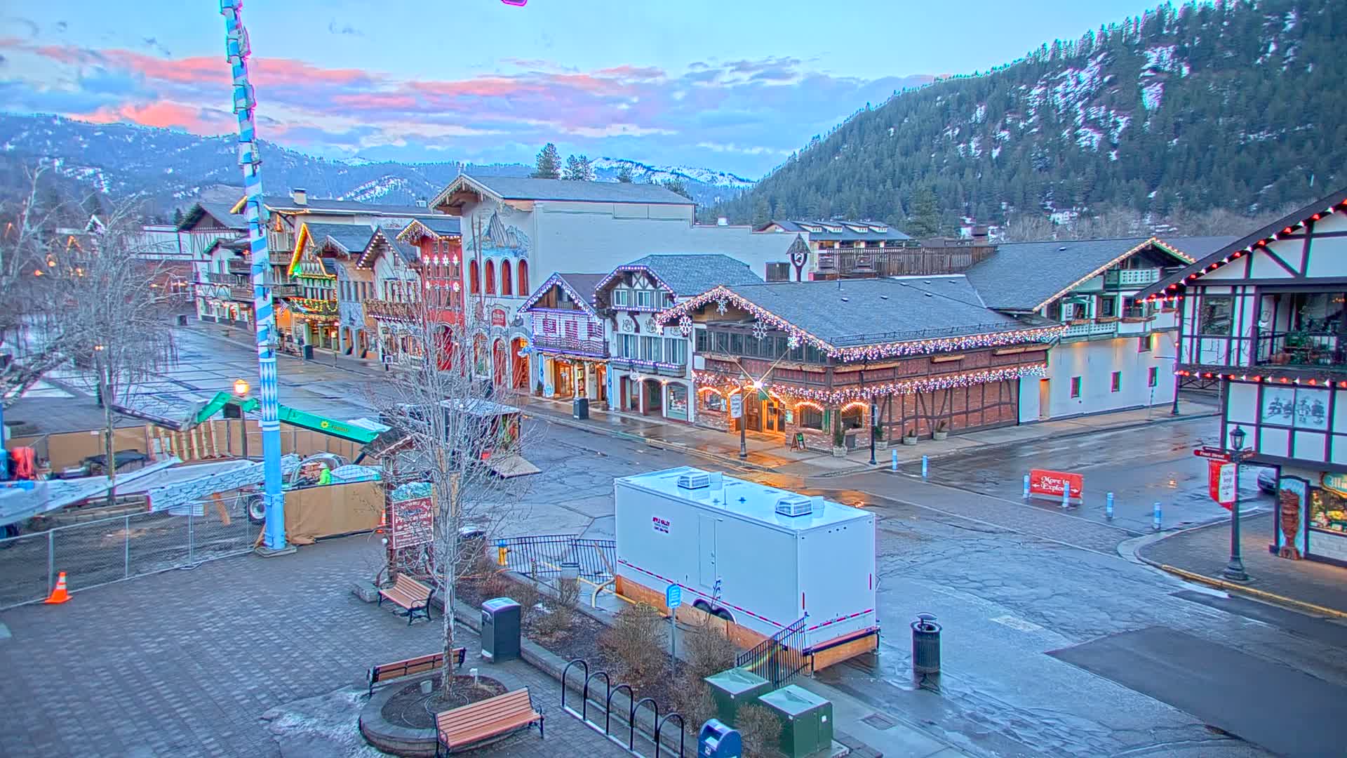 A town square with Bavarian-style buildings decorated with lights, under a twilight sky with a mountain range in the background.