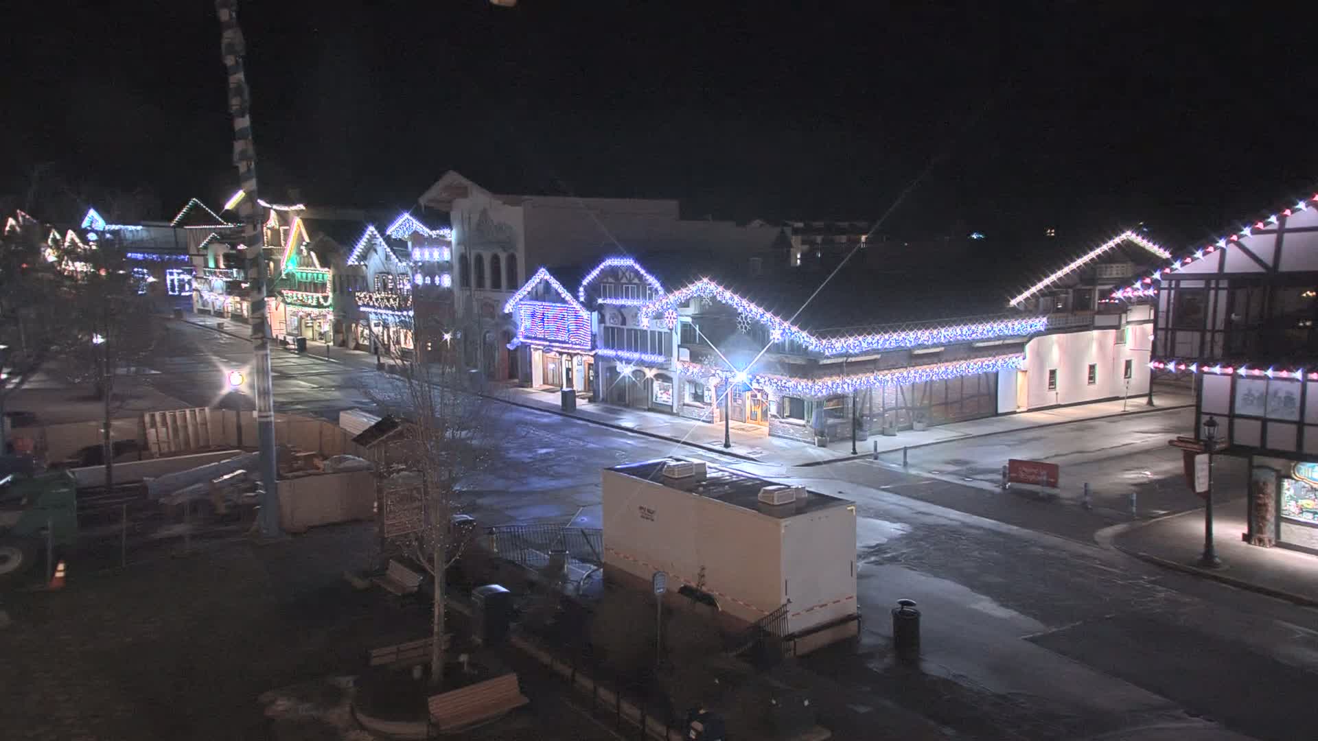 A nighttime, snow-dusted street scene shows a row of buildings decorated with strings of holiday lights.