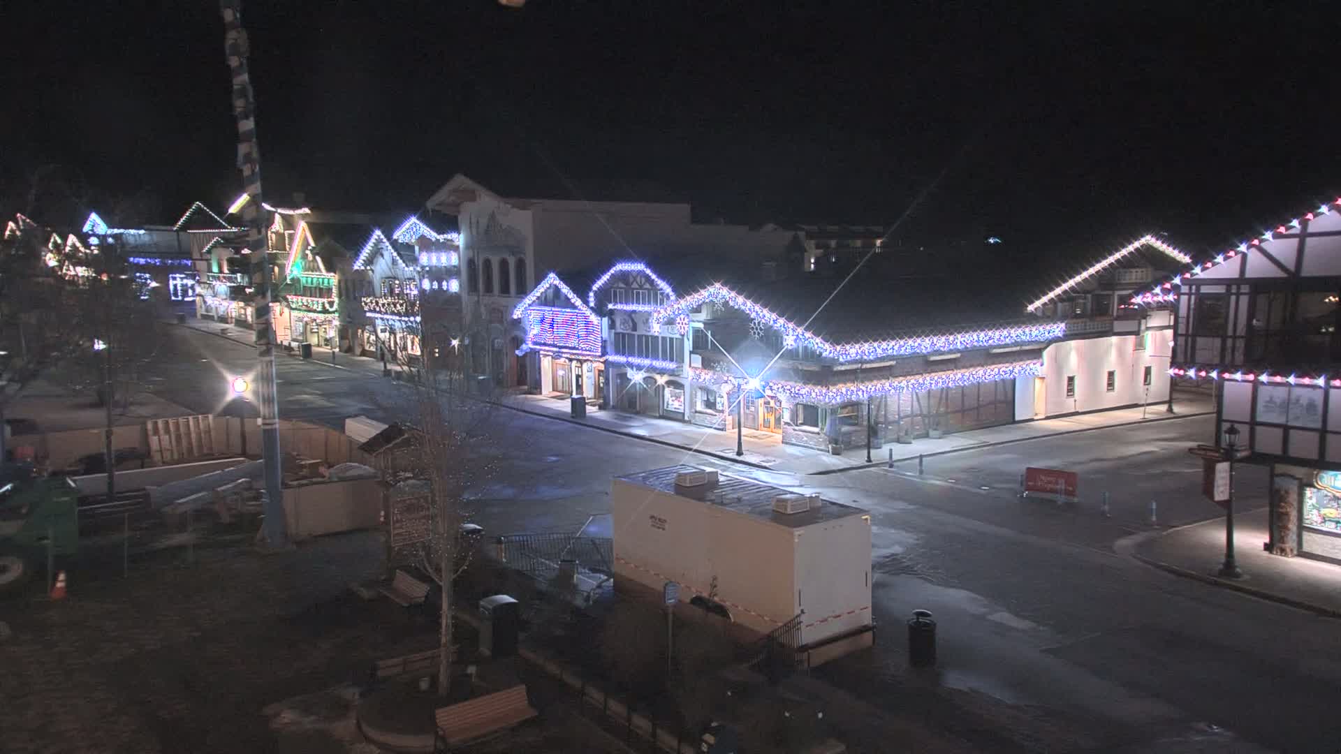 A nighttime street scene shows a row of buildings decorated with twinkling lights, a mostly empty street, and a few parked vehicles.