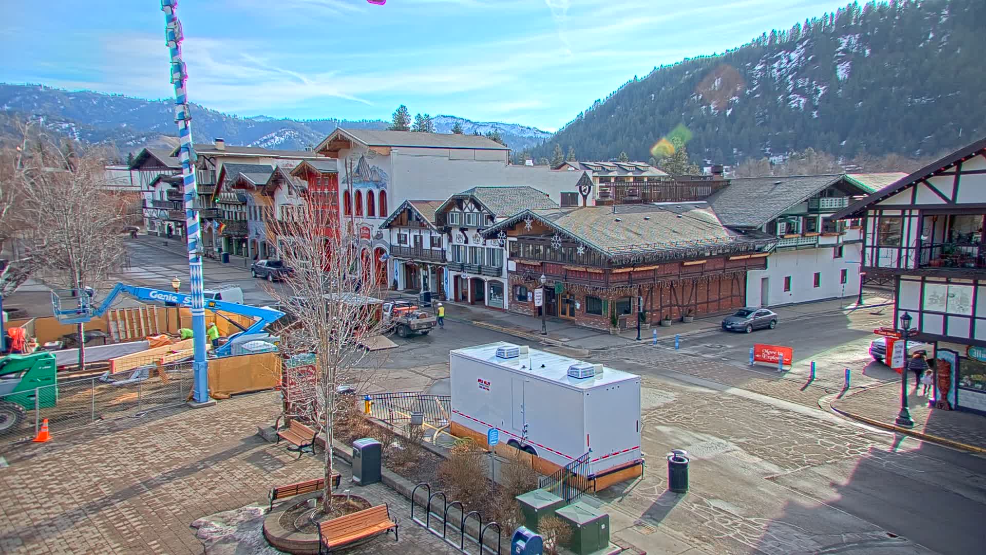 A town square with Bavarian-style buildings under a sunny sky, surrounded by snow-capped mountains, features construction equipment and workers.
