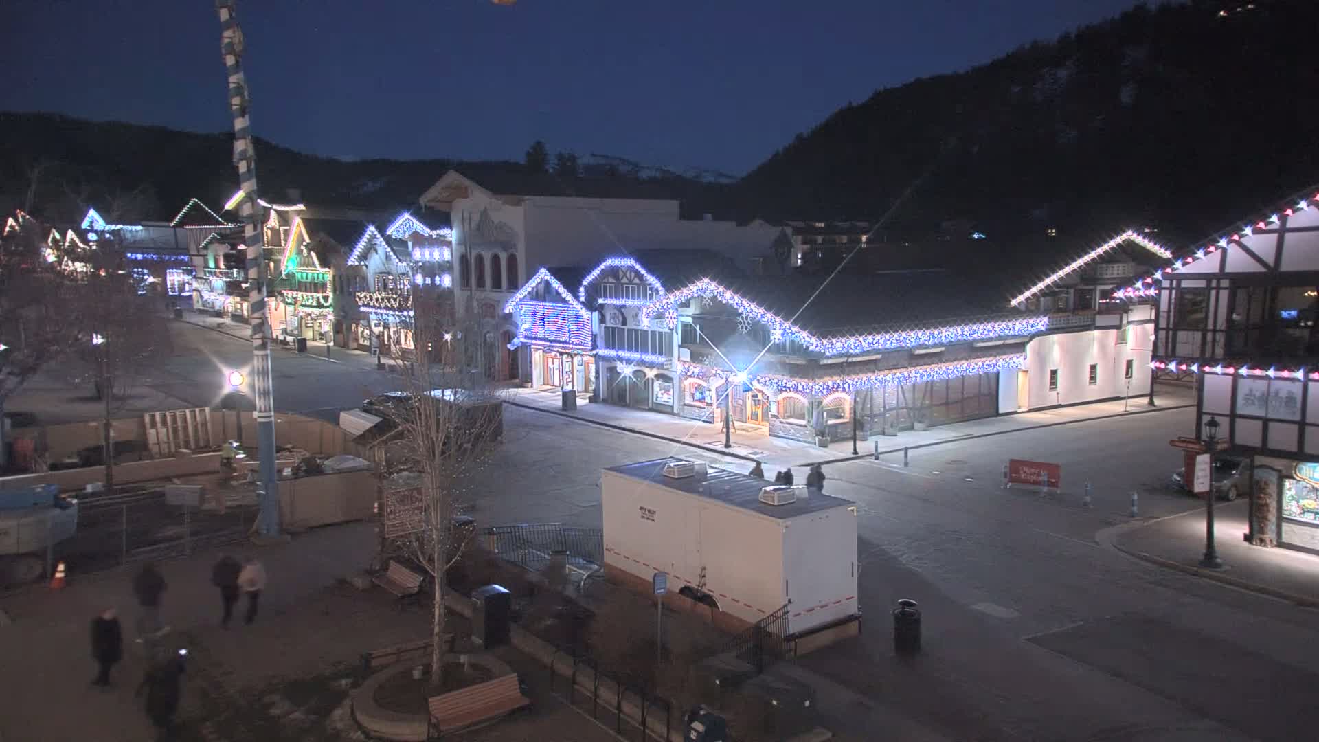 A nighttime, snow-covered street scene shows a row of buildings decorated with Christmas lights, and a few people walking along the street.