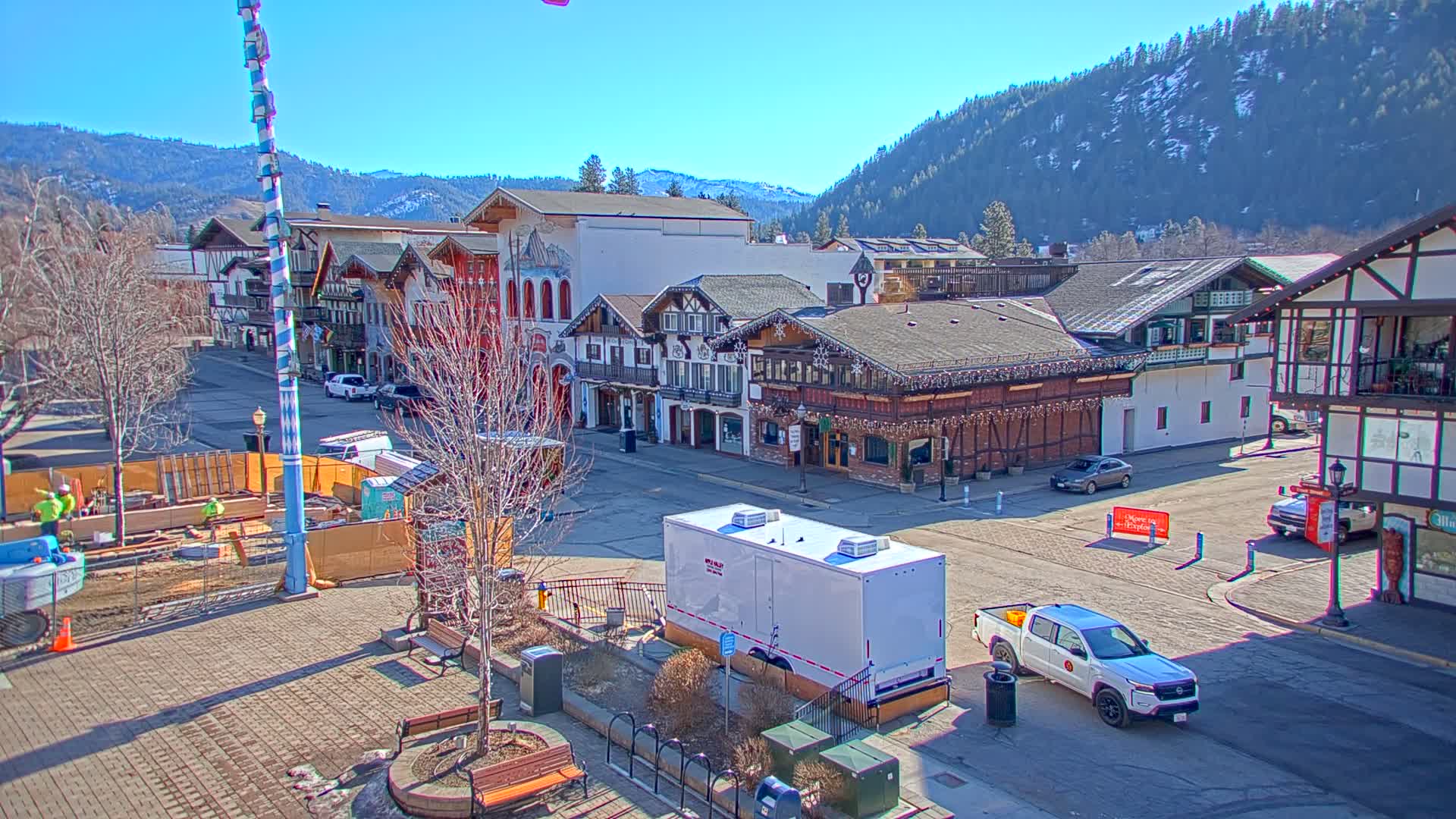 A sunny day reveals a town square with Bavarian-style buildings, construction underway, and vehicles parked on a street against a backdrop of snow-capped mountains.