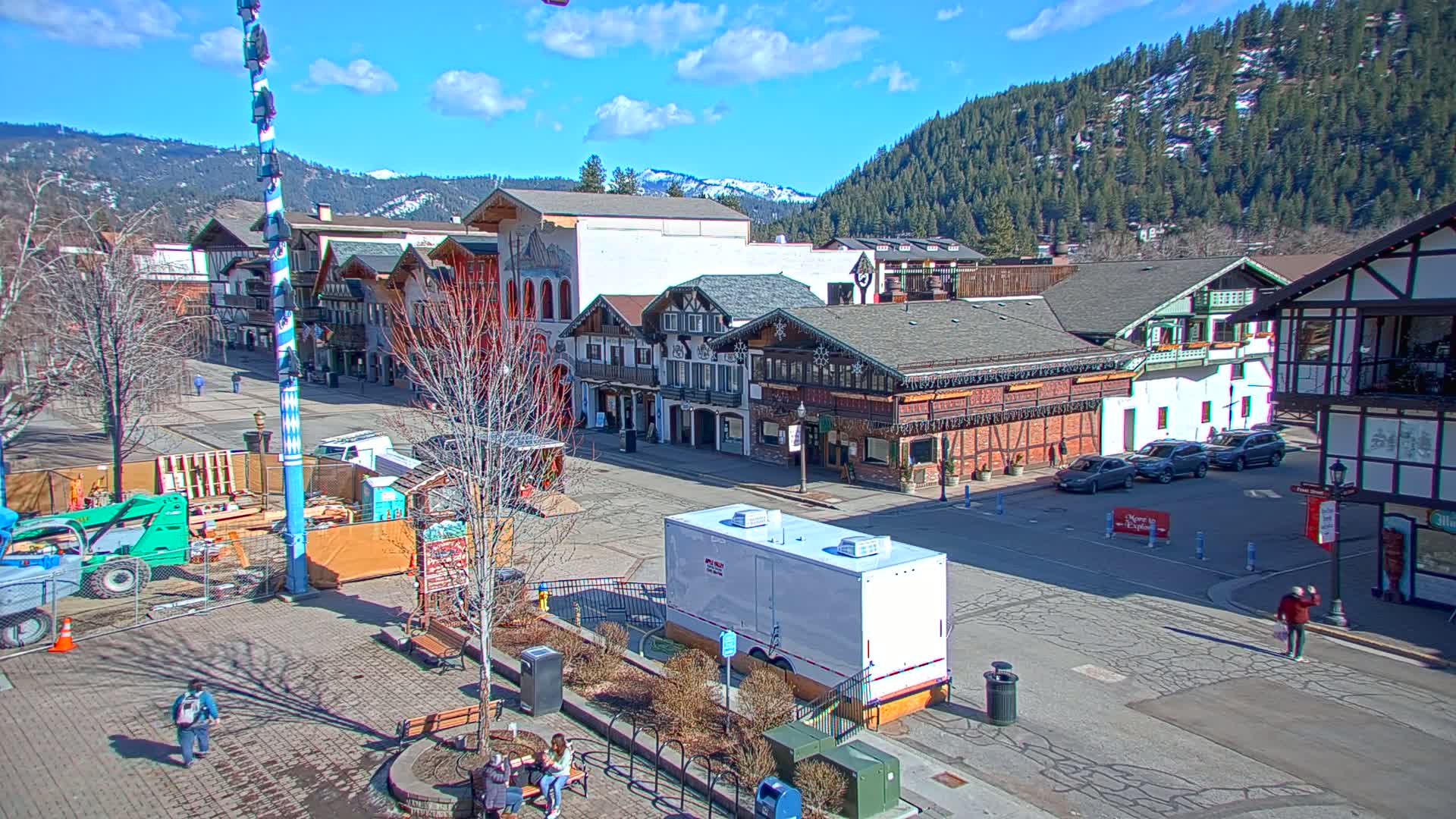 A sunny day reveals a town square with European-style buildings, construction equipment, and a few pedestrians, all set against a backdrop of snow-capped mountains and evergreen trees.