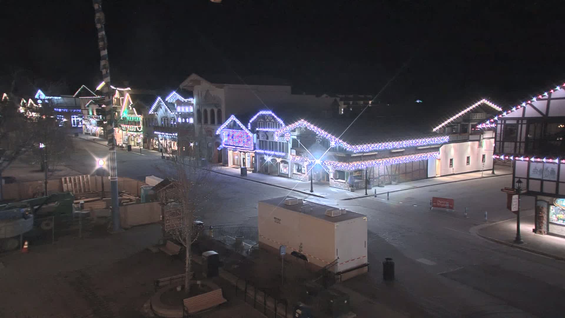 A nighttime view of a town square with several buildings decorated with twinkling lights, some construction equipment, and a portable restroom.