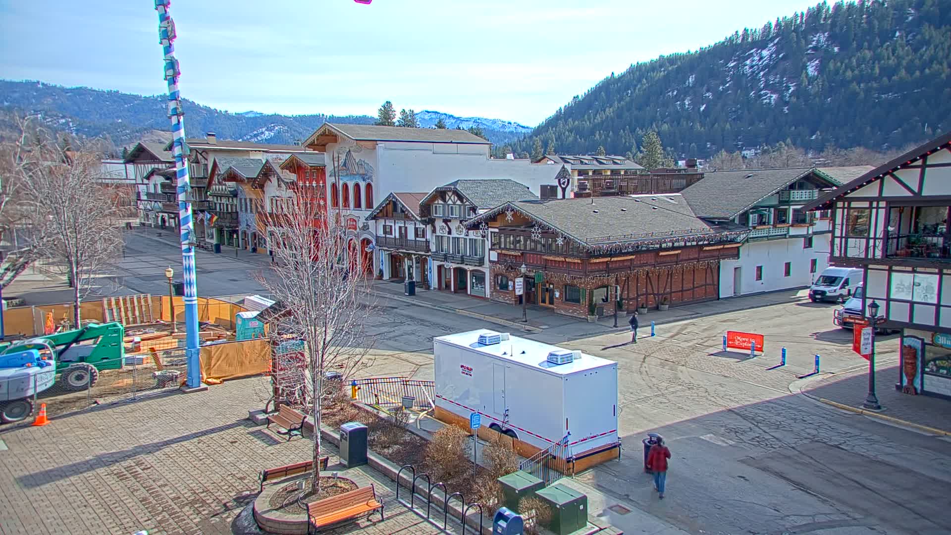 A town square with Bavarian-style buildings under a sunny sky, surrounded by snow-capped mountains in the background, shows construction equipment and a portable restroom.