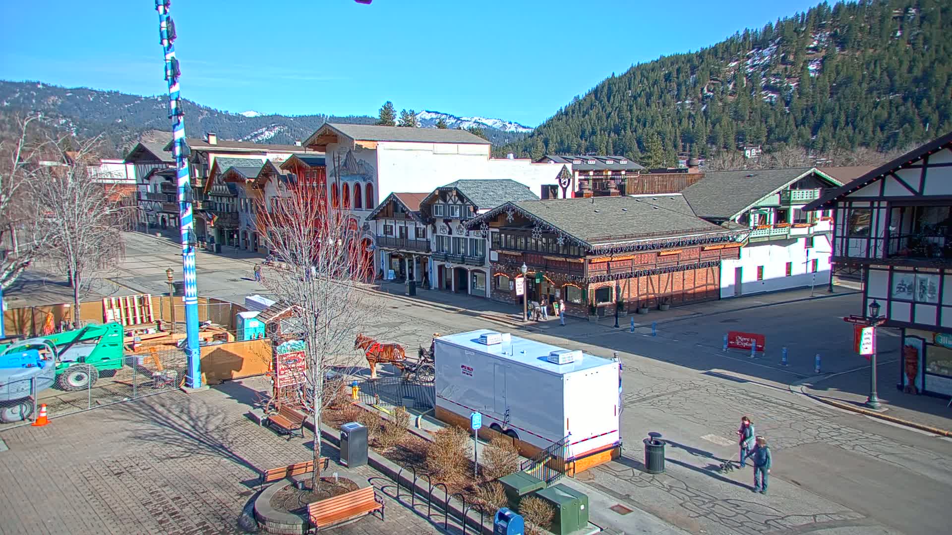 A sunny day reveals a quaint town square with Bavarian-style buildings, a horse-drawn carriage, and construction underway, all nestled against a backdrop of snow-capped mountains and evergreen forests.