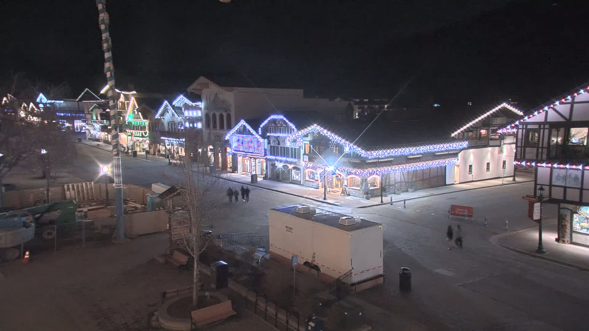 A nighttime view of a town square with festively lit buildings and a few pedestrians, under clear skies.