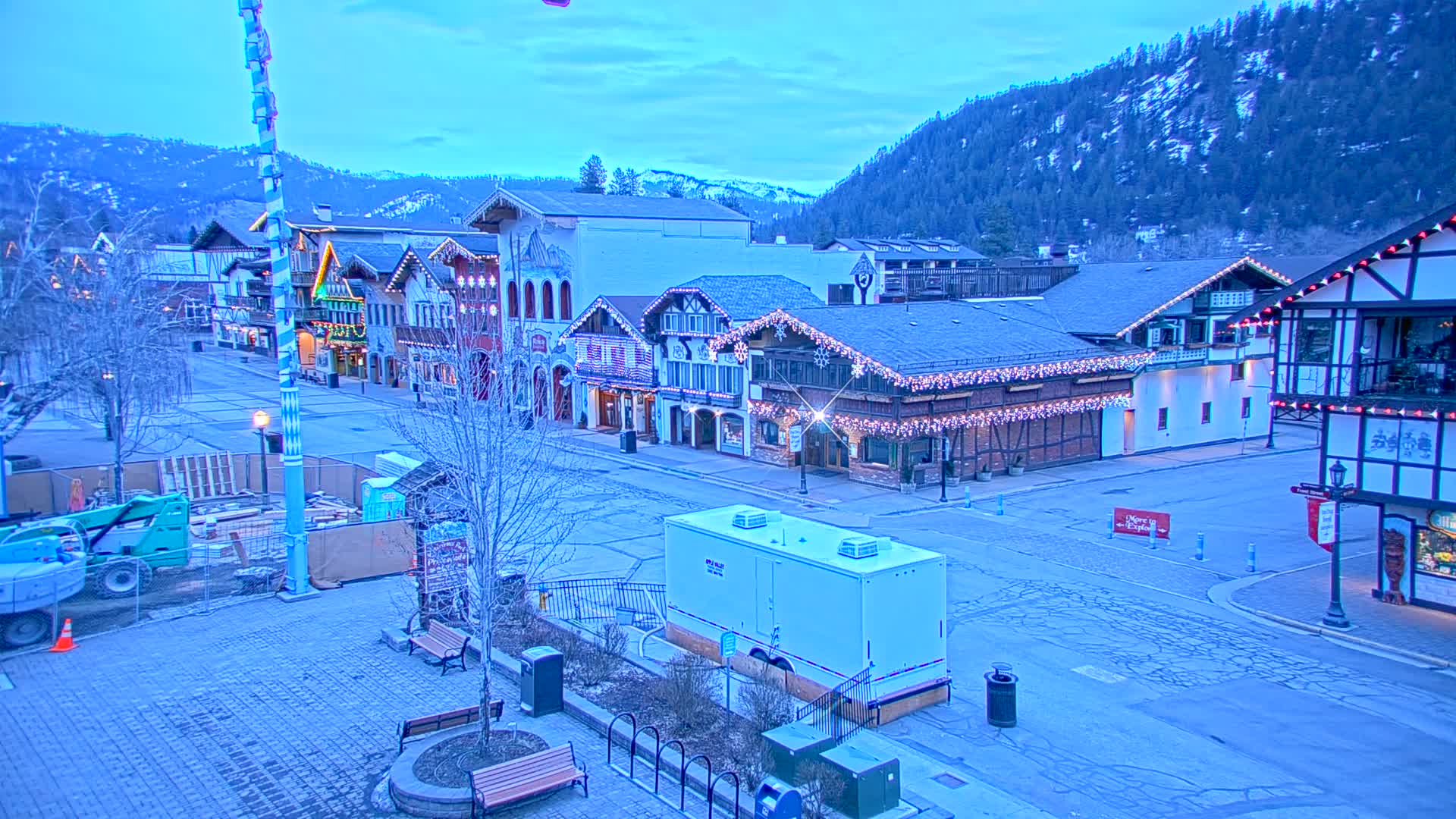 A mostly empty town square with Bavarian-style buildings decorated with string lights, under a twilight sky, with a mountain in the background and construction equipment present.
