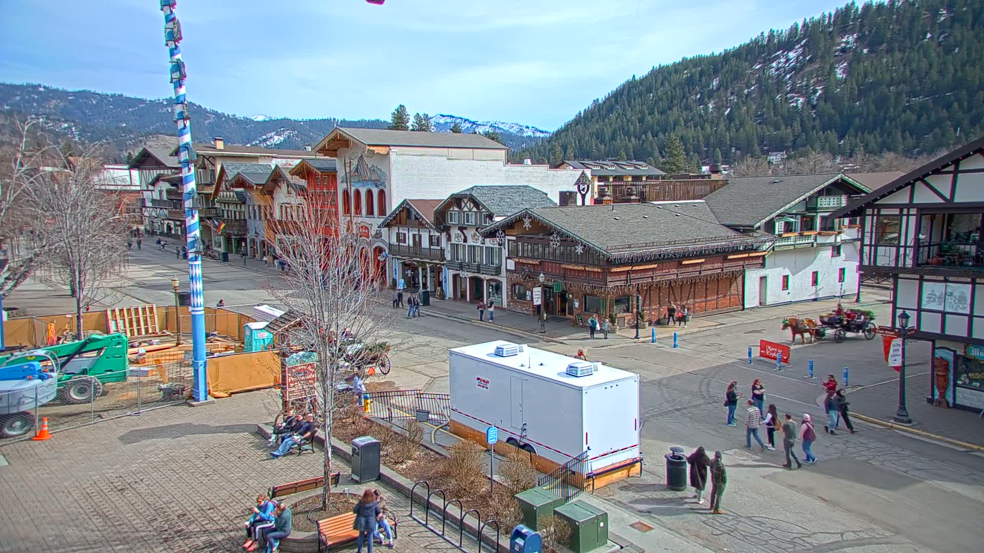 A town square with Bavarian-style buildings, a horse-drawn carriage, and several people walking around on a sunny day with snow-capped mountains in the background.