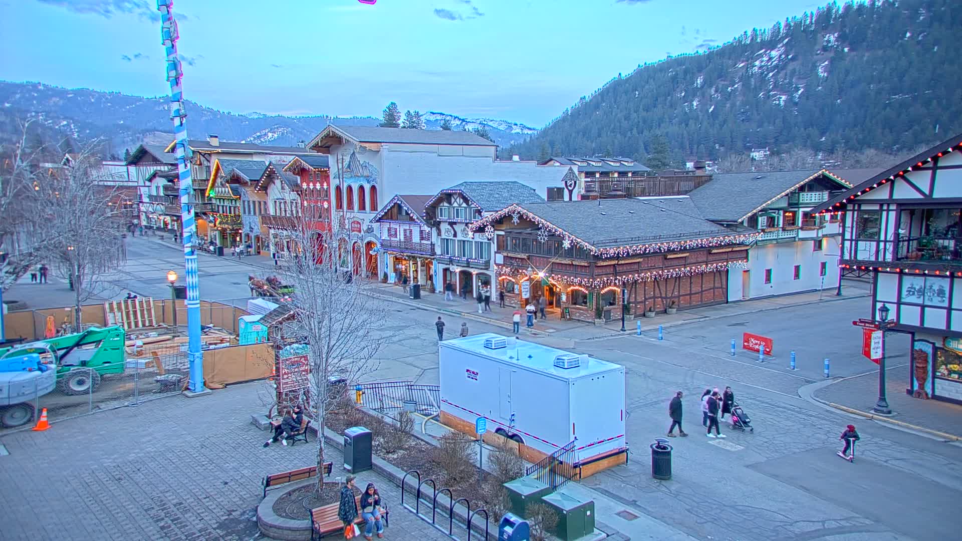 A town square with Bavarian-style buildings under a twilight sky, featuring a few pedestrians and construction equipment.