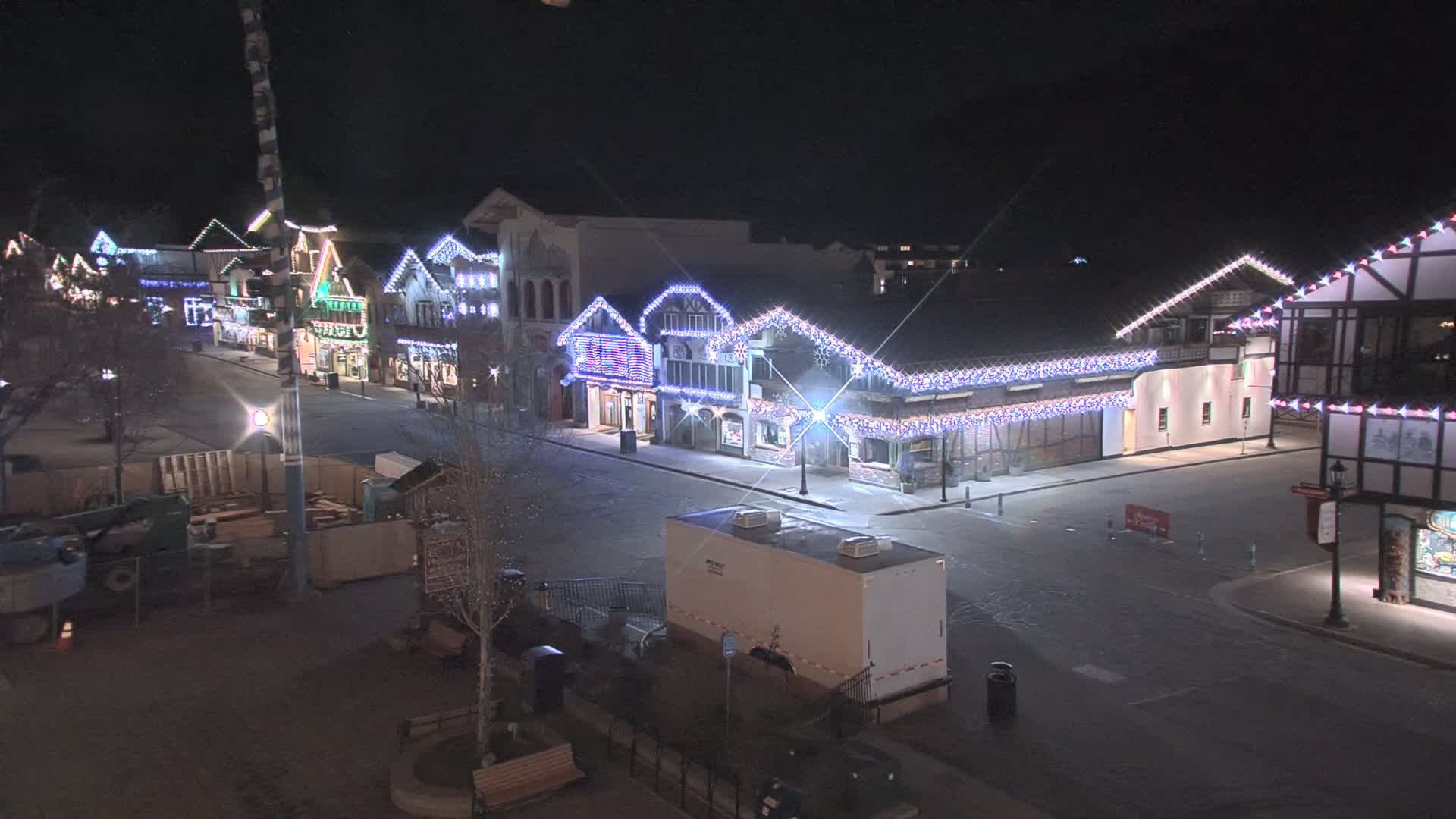 A mostly empty street at night lined with festively decorated buildings, lightly snow-covered.