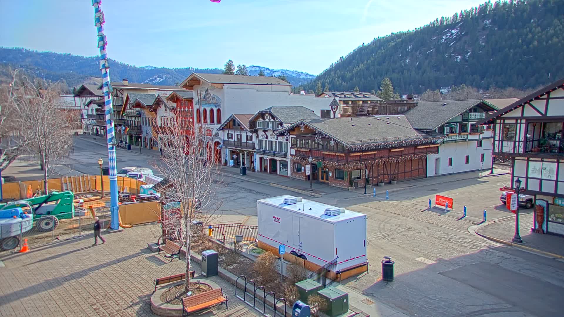 A mostly empty town square with Bavarian-style buildings under a clear, sunny sky, backed by snow-capped mountains.
