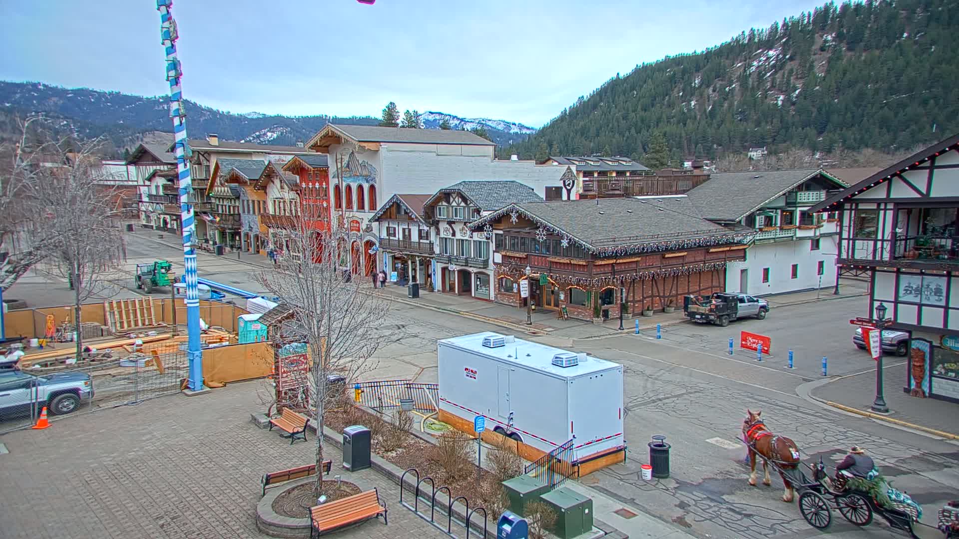 A horse-drawn carriage waits on a paved street in front of a row of Bavarian-style buildings situated at the base of a snow-dusted mountain range under a cloudy sky.