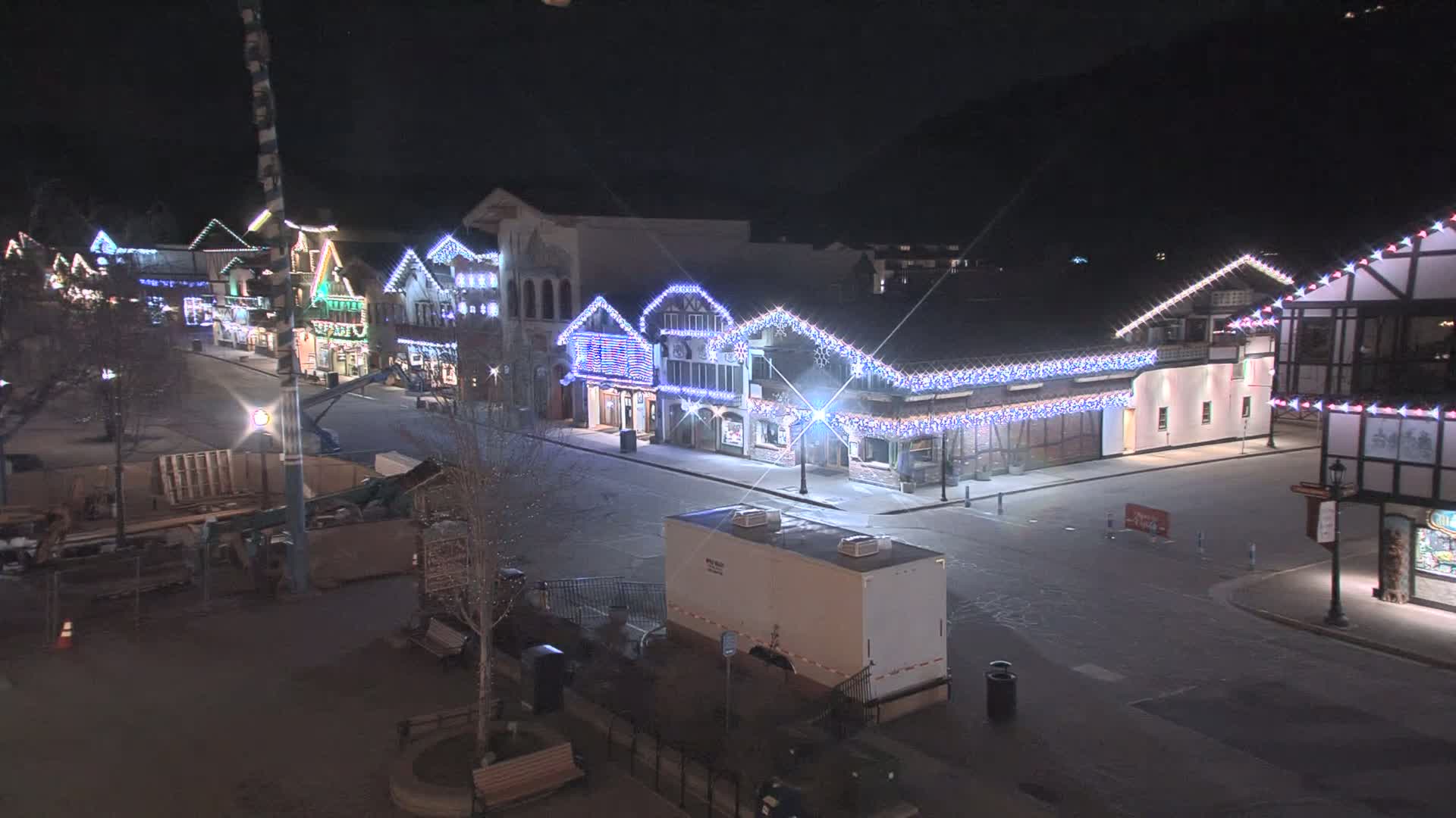 A mostly empty street at night, lined with festively lit buildings, shows some construction equipment and a portable restroom; the weather appears clear.