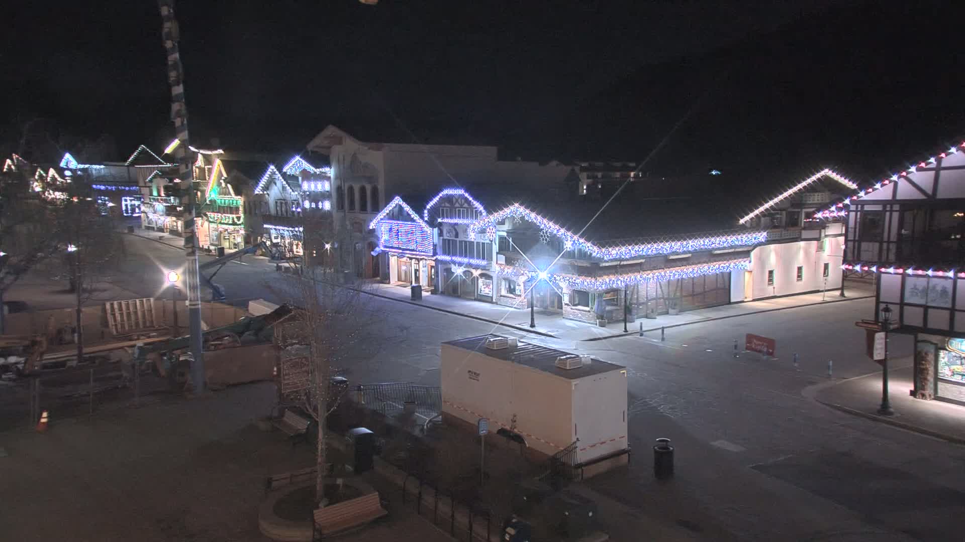 A mostly empty street at night lined with buildings decorated with twinkling lights, possibly snow on the ground.