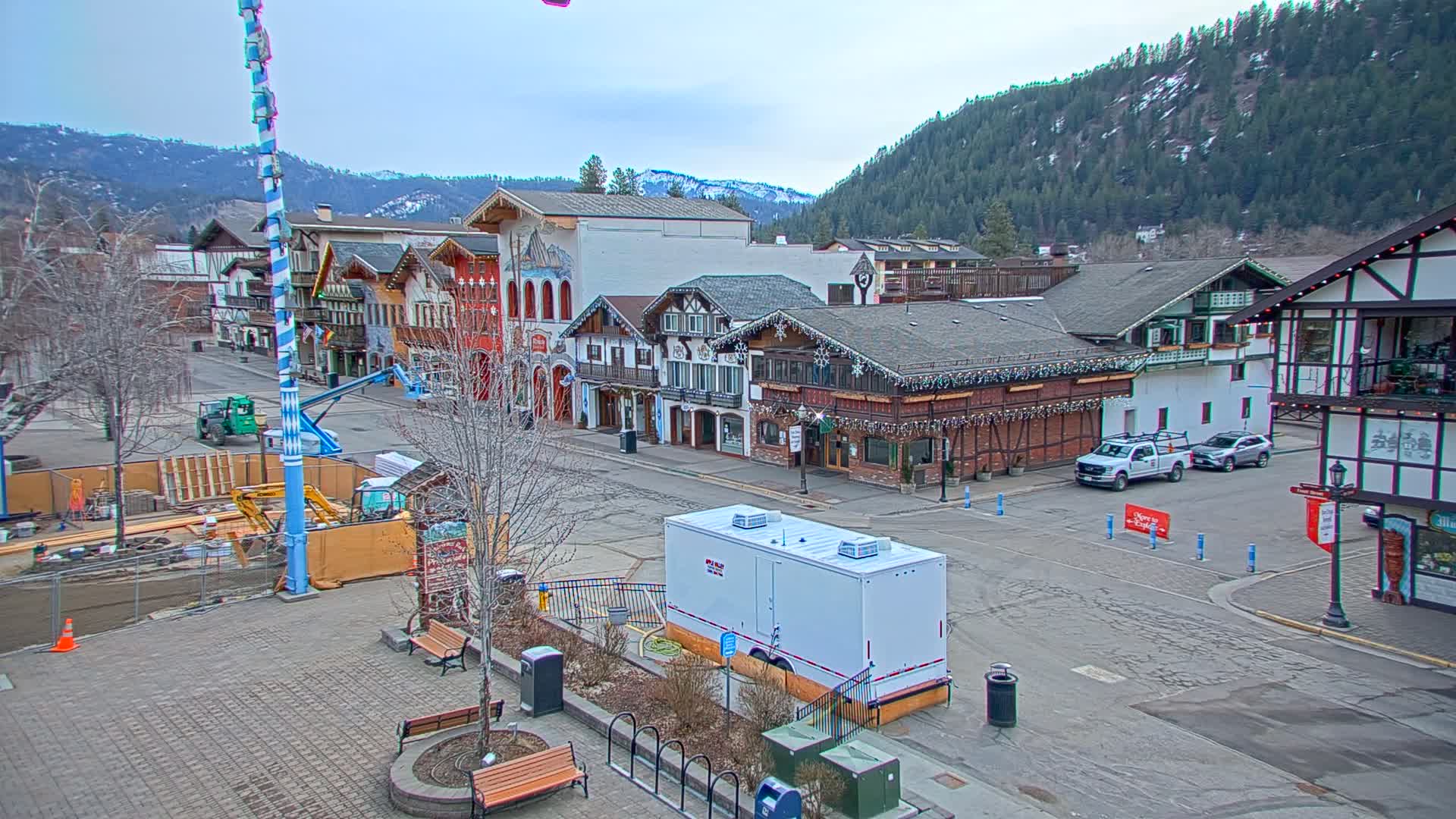 A town square with several European-style buildings under a cloudy sky, construction equipment, and a large white trailer are visible, with a mountain range in the background.