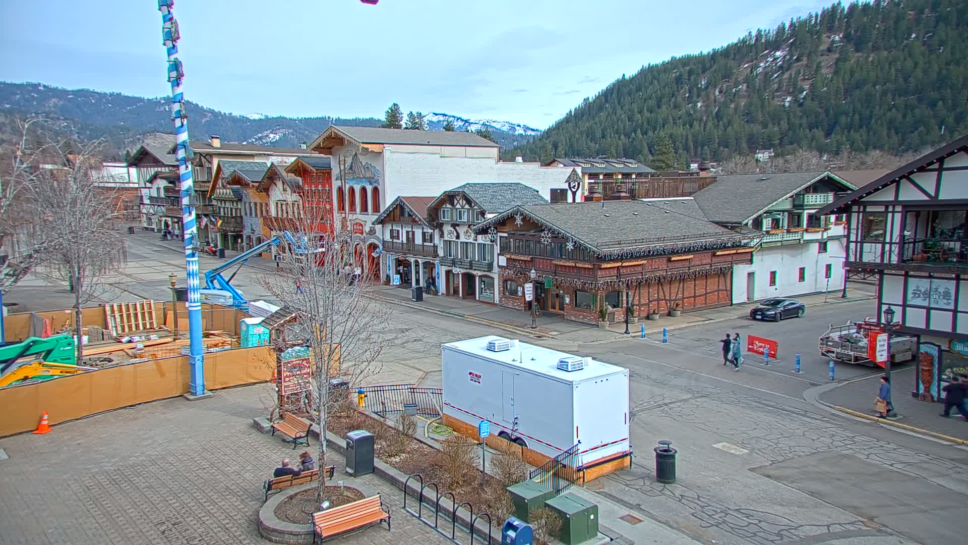 A town square with Bavarian-style buildings, under a mostly clear sky, with mountains in the background and construction underway.