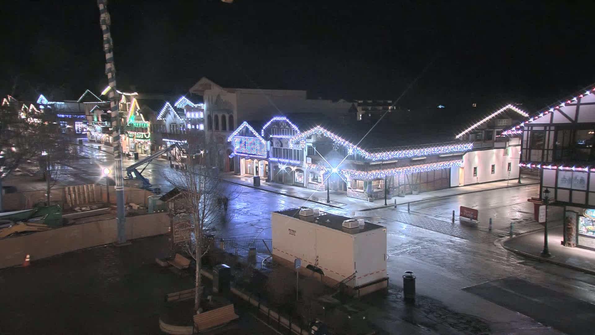 A nighttime view of a town square with buildings decorated with twinkling lights, under a dark sky and wet streets.