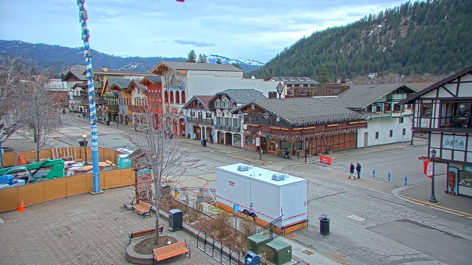 A town square with several Bavarian-style buildings, under a mostly cloudy sky, features a construction site and a few pedestrians.