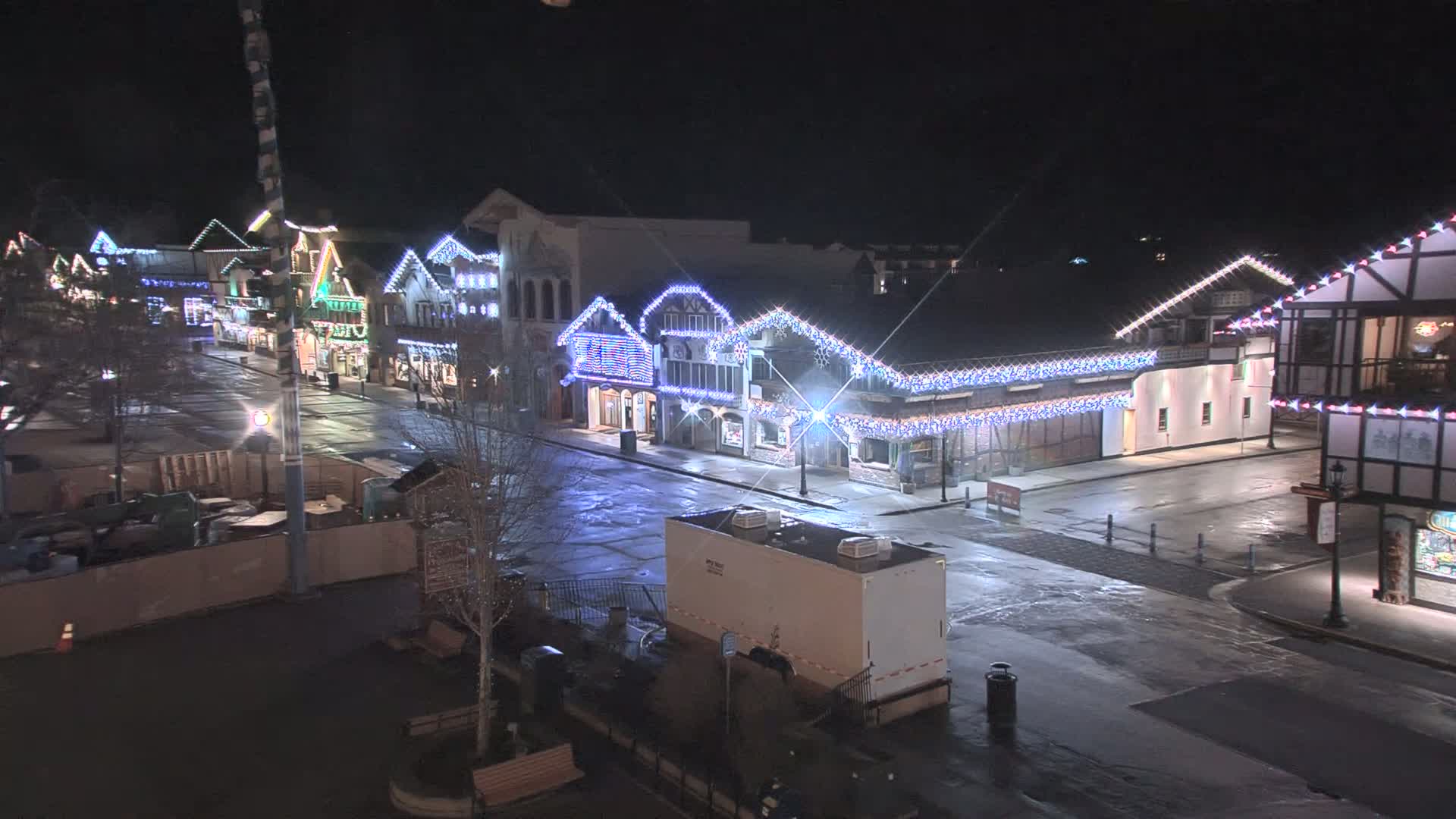 A nighttime street scene shows a row of buildings decorated with strings of lights, a mostly empty street, and a portable restroom.