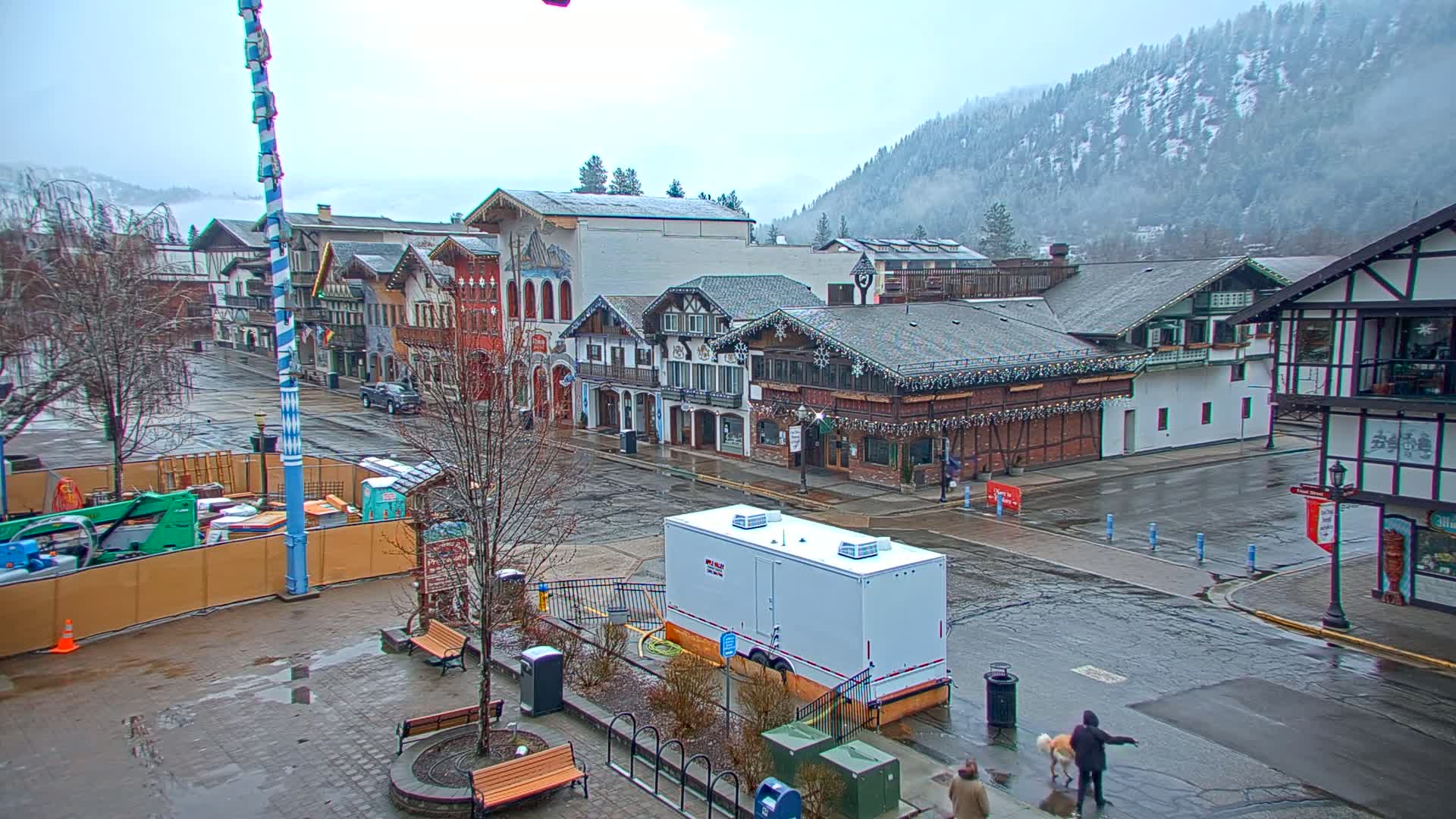 A wet, overcast town square with several multi-story buildings in a European style, a snow-covered mountain in the background, and a person walking a dog in the foreground.