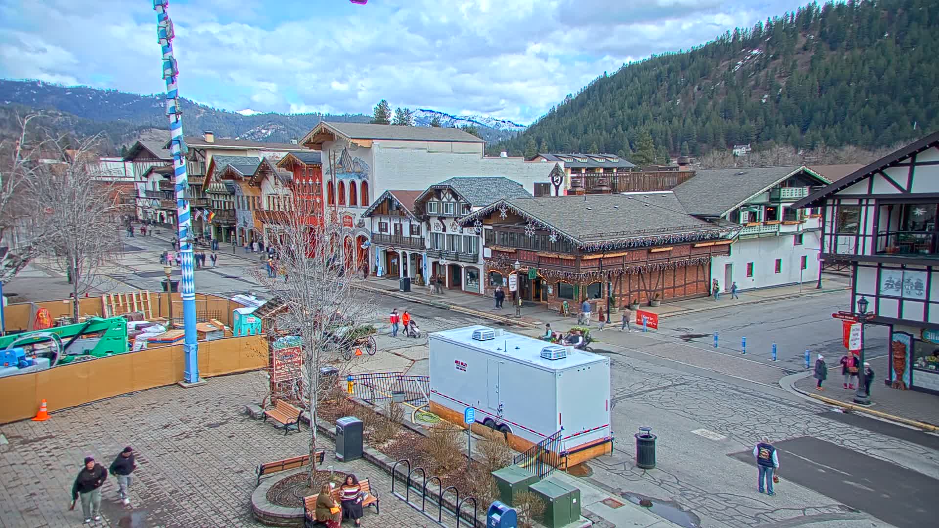 A partly cloudy day reveals a town square with several Bavarian-style buildings, a few pedestrians, and construction equipment.