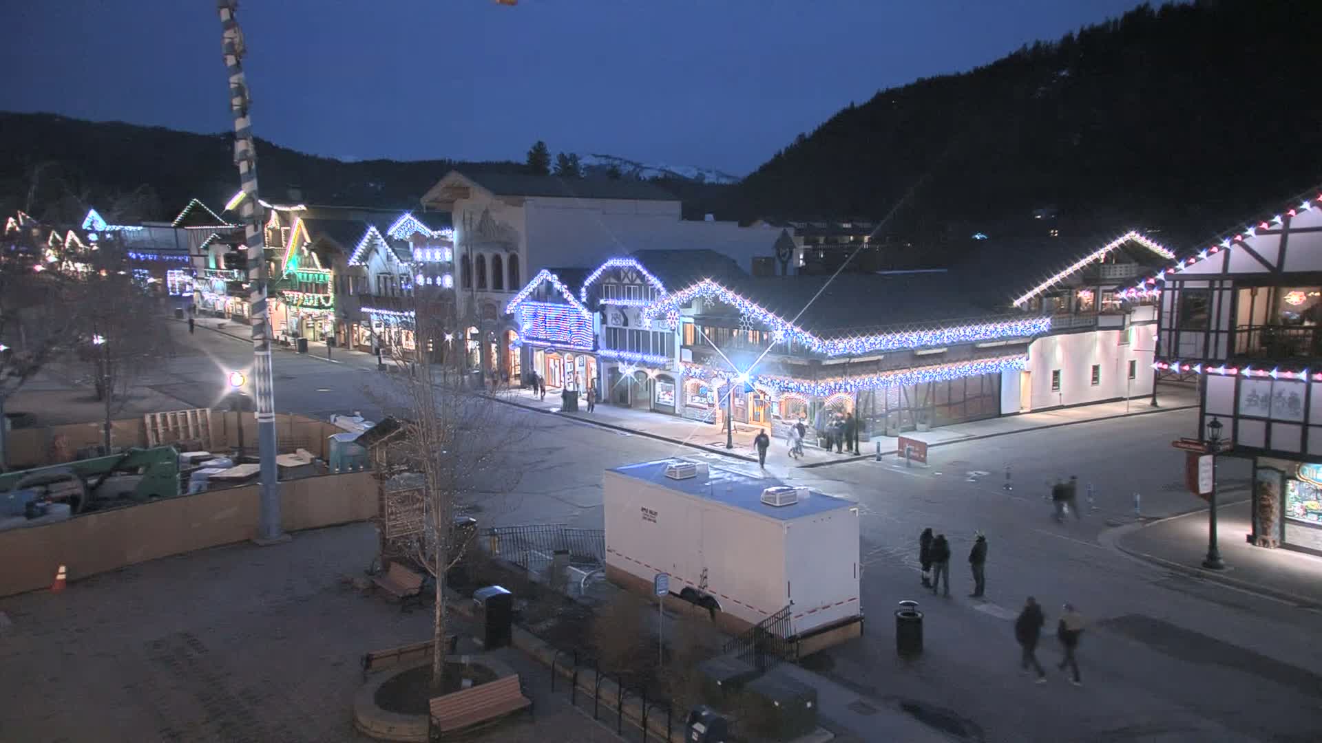 A nighttime view of a town square with buildings decorated with Christmas lights, under a clear, dark sky.