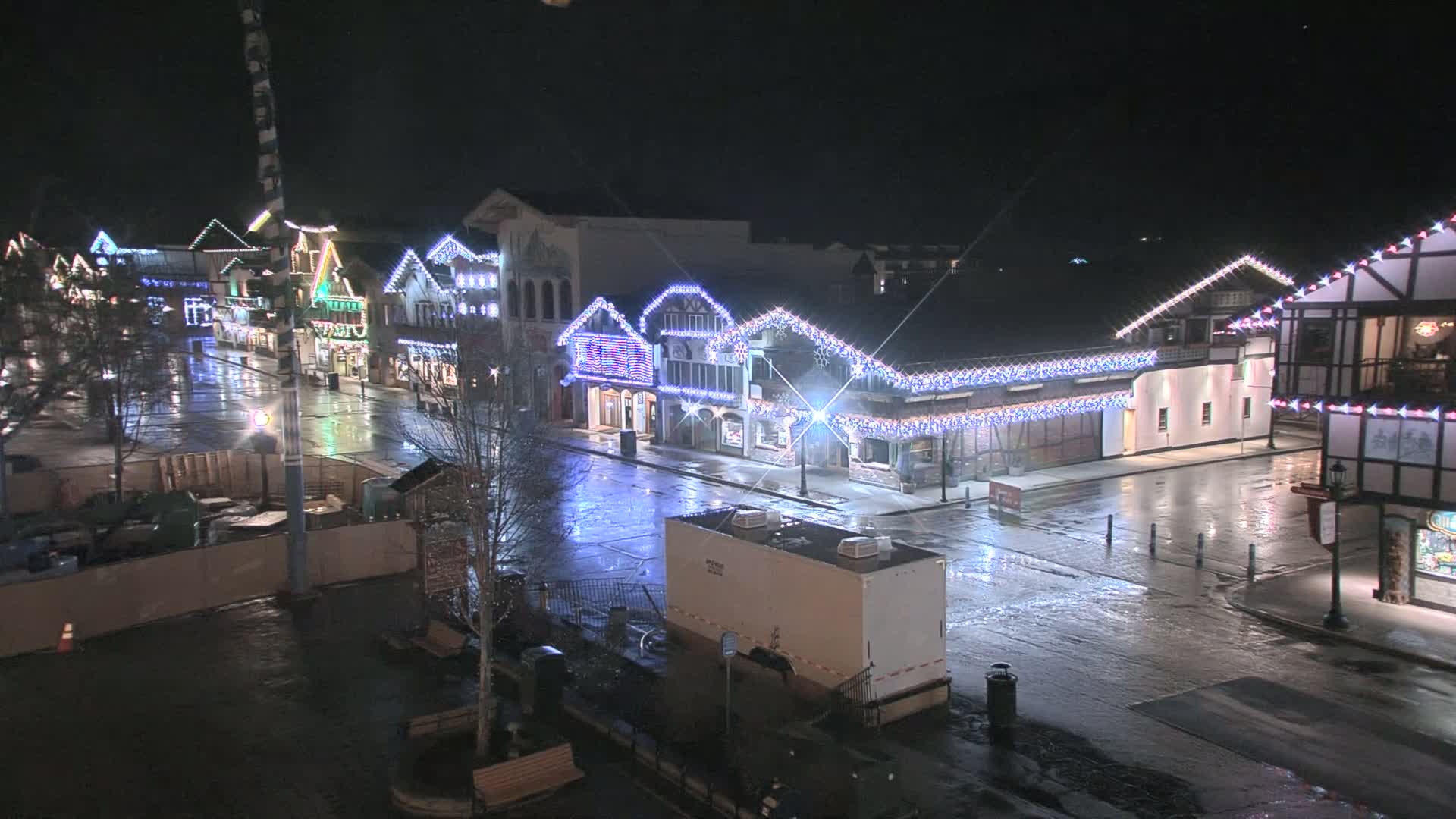 A rain-slicked town square at night, illuminated by strings of lights on the buildings.