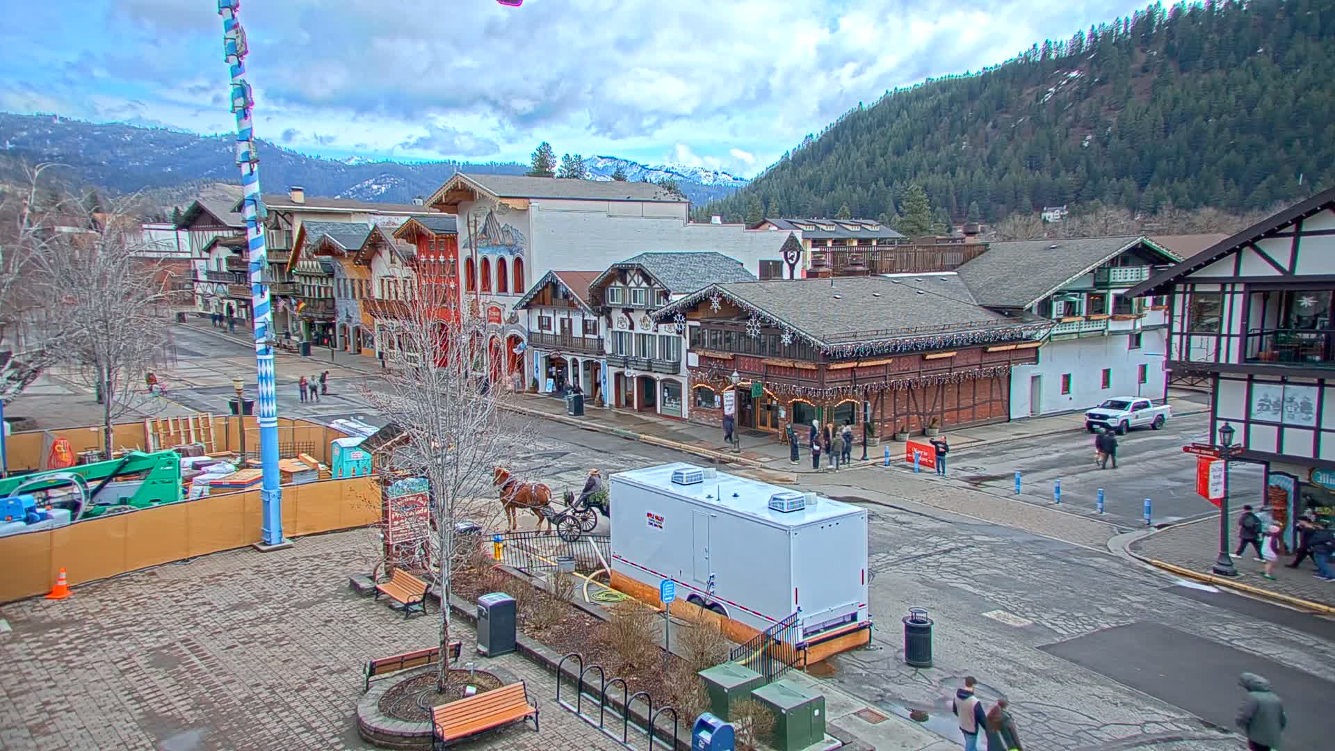 A horse-drawn carriage is on a street lined with Bavarian-style buildings under a cloudy sky, with mountains in the background.