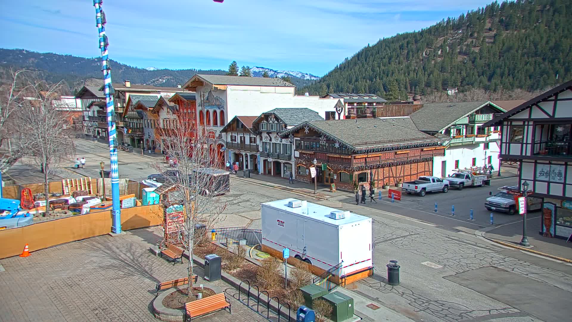 A mostly sunny day reveals a town square with Bavarian-style buildings, a few parked vehicles, and construction or event preparation underway.