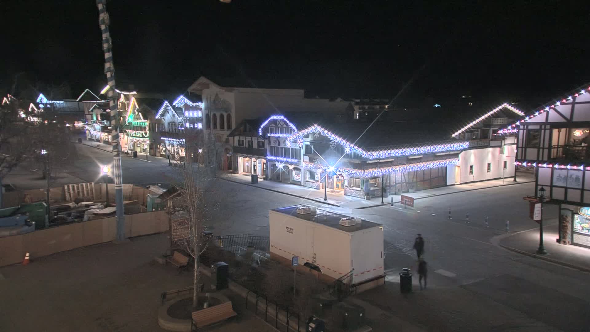 A nighttime, empty street lined with festively lit buildings, and a few people walking.