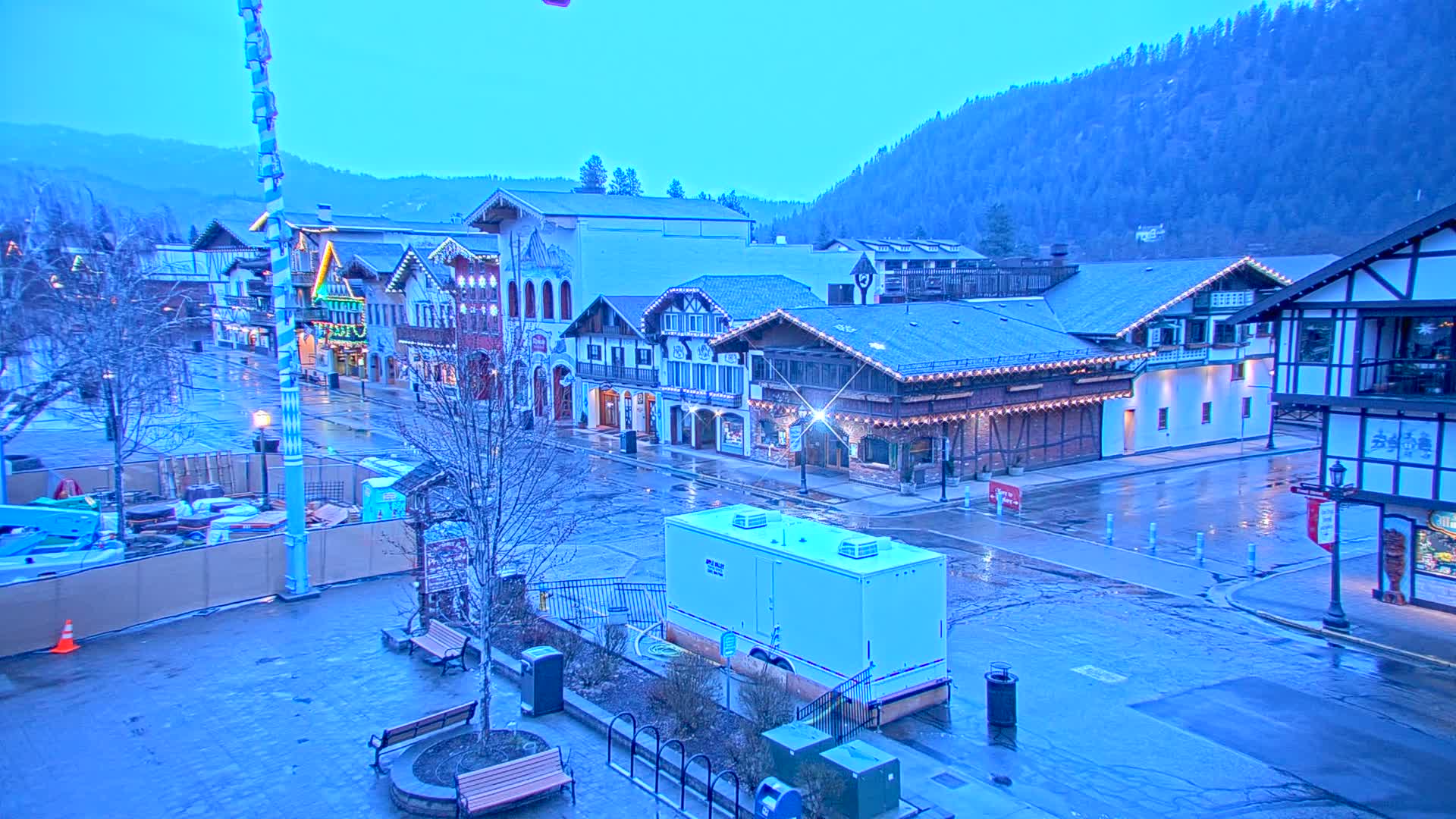 A wet, empty town square with Bavarian-style buildings under a twilight sky, backed by a mountain range.