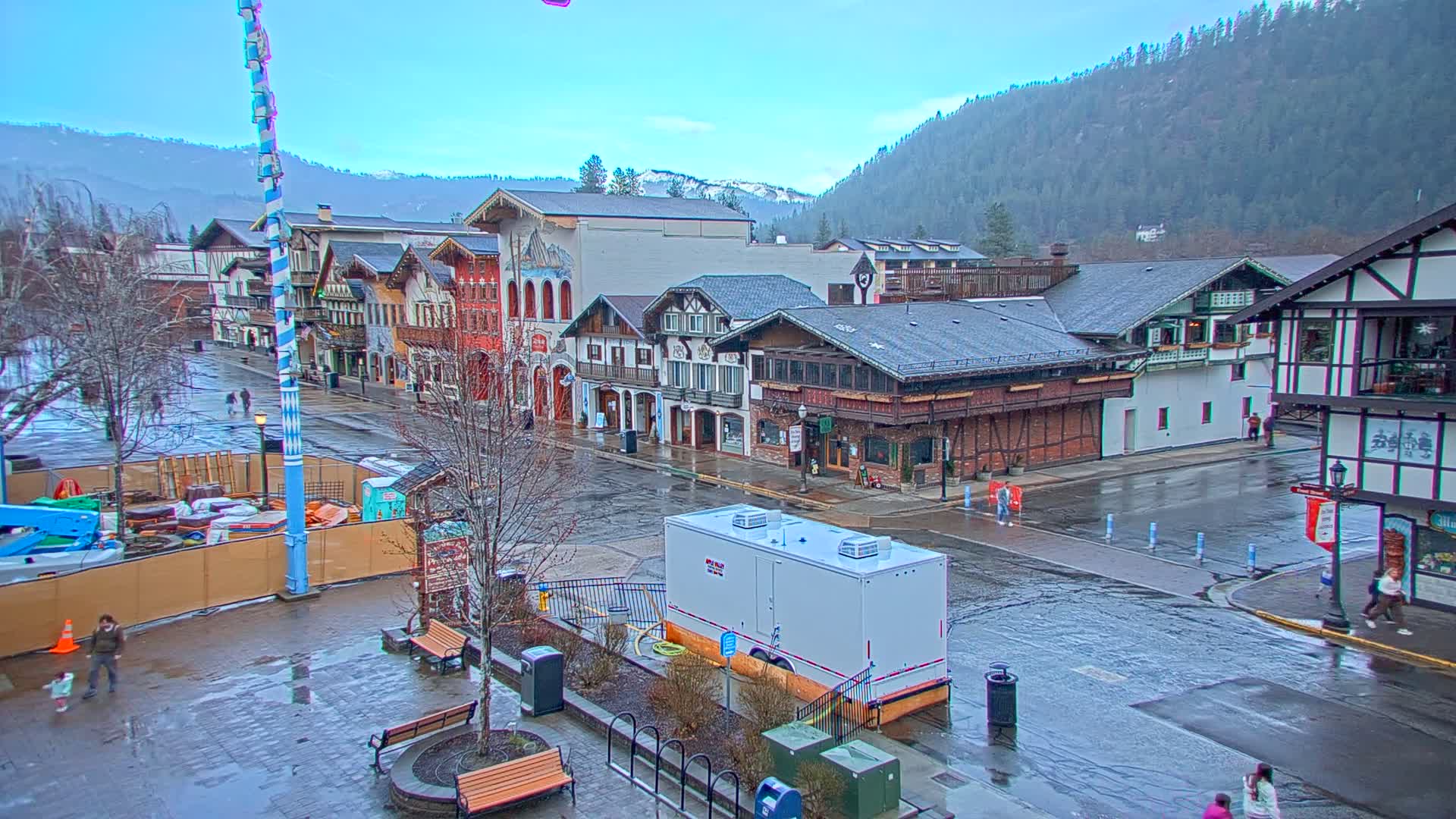 A rain-soaked town square with Bavarian-style buildings, a tall maypole, and a few pedestrians is nestled against a backdrop of a mountain range under an overcast sky.