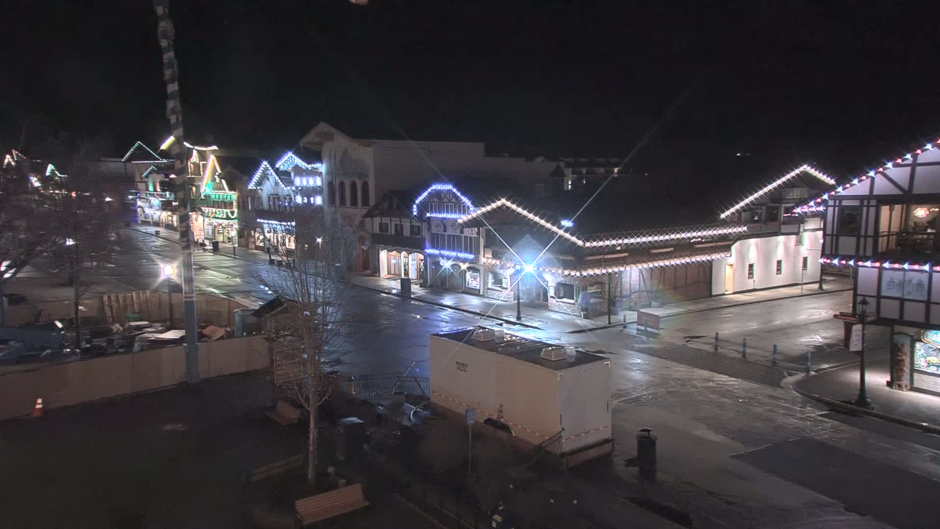A nighttime, wet street scene shows a row of festively lit buildings with a few parked vehicles and construction barriers.