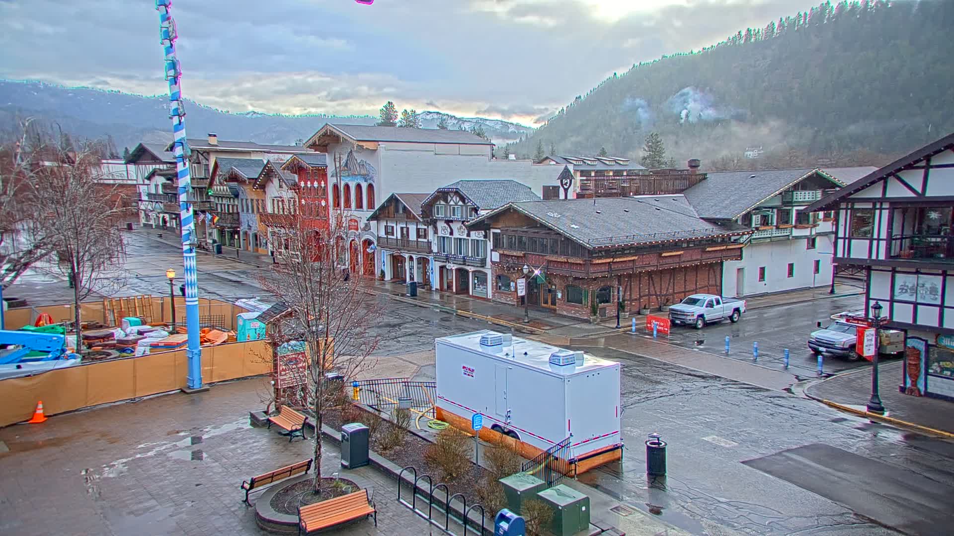 A wet town square with Bavarian-style buildings, a tall maypole, and mountains in the background under an overcast sky.