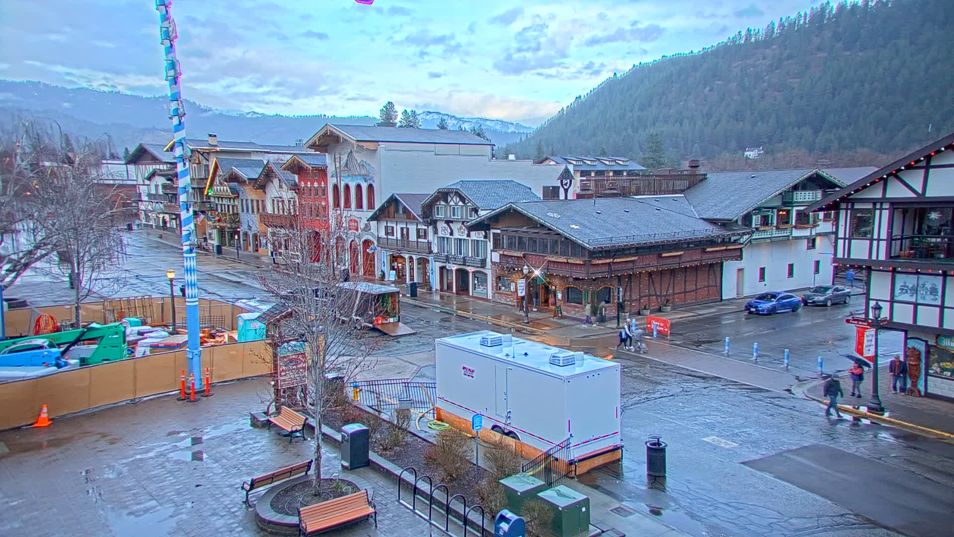 A rain-slicked town square with Bavarian-style buildings, a few pedestrians, and some vehicles is nestled in a valley beneath a mountain range under a cloudy sky.