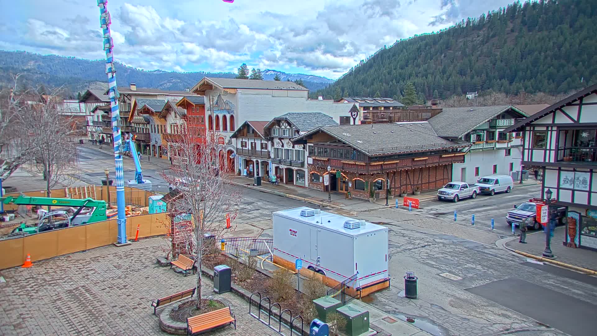 A town square with Bavarian-style buildings, construction underway, and a partly cloudy sky, nestled against a backdrop of forested mountains.