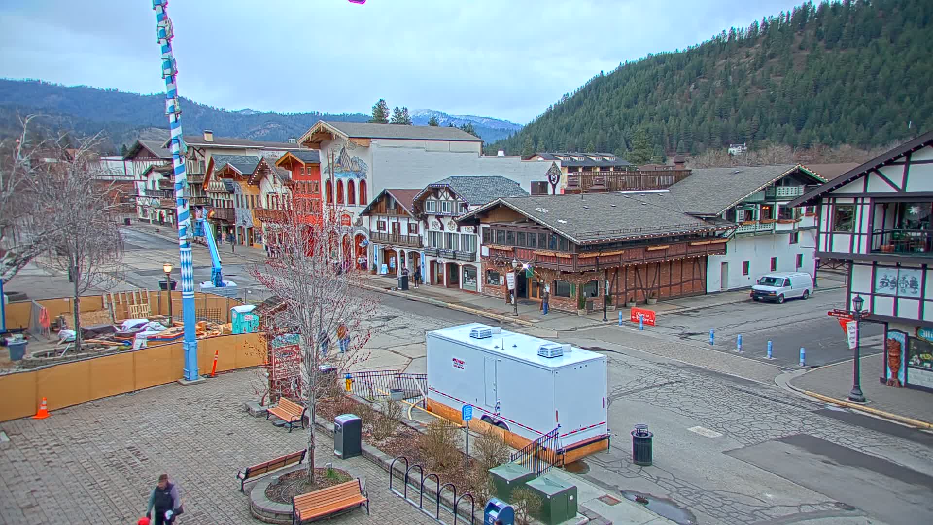A town square with Bavarian-style buildings under an overcast sky, featuring a construction area and a portable restroom.
