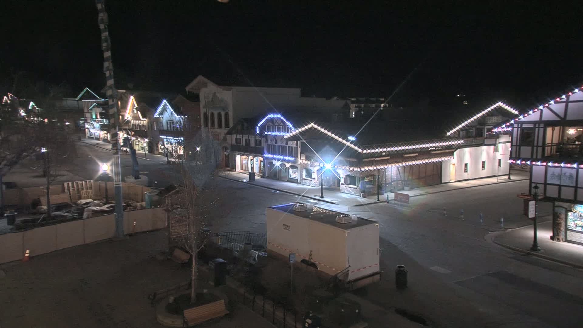 A nighttime, empty street lined with festively lit buildings, and a portable restroom sits near a construction site.