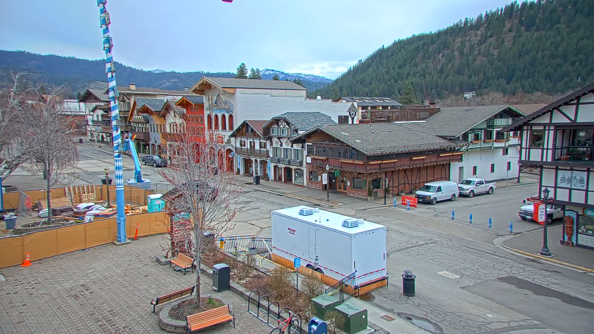 A town square with Bavarian-style buildings, bare trees, and a few parked vehicles under an overcast sky.
