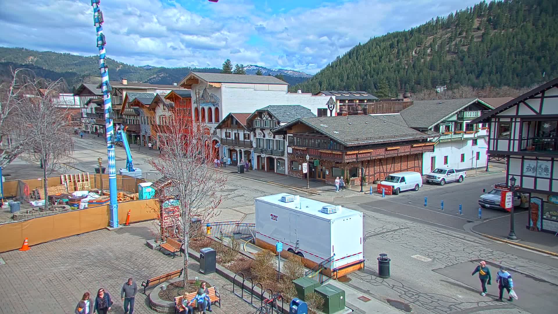 A partly cloudy day reveals a town square with several Bavarian-style buildings, a tall maypole, and some people walking around.