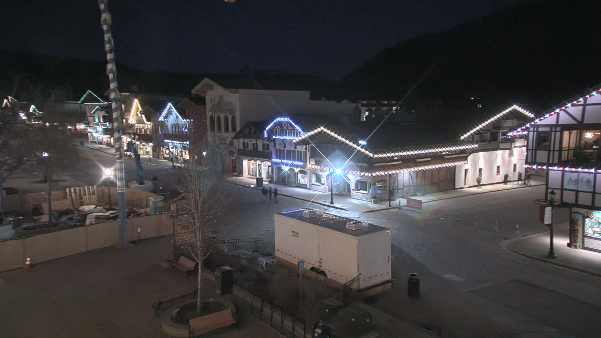 A nighttime street scene shows a row of buildings decorated with string lights, a mostly empty street, and a construction site behind a fence.