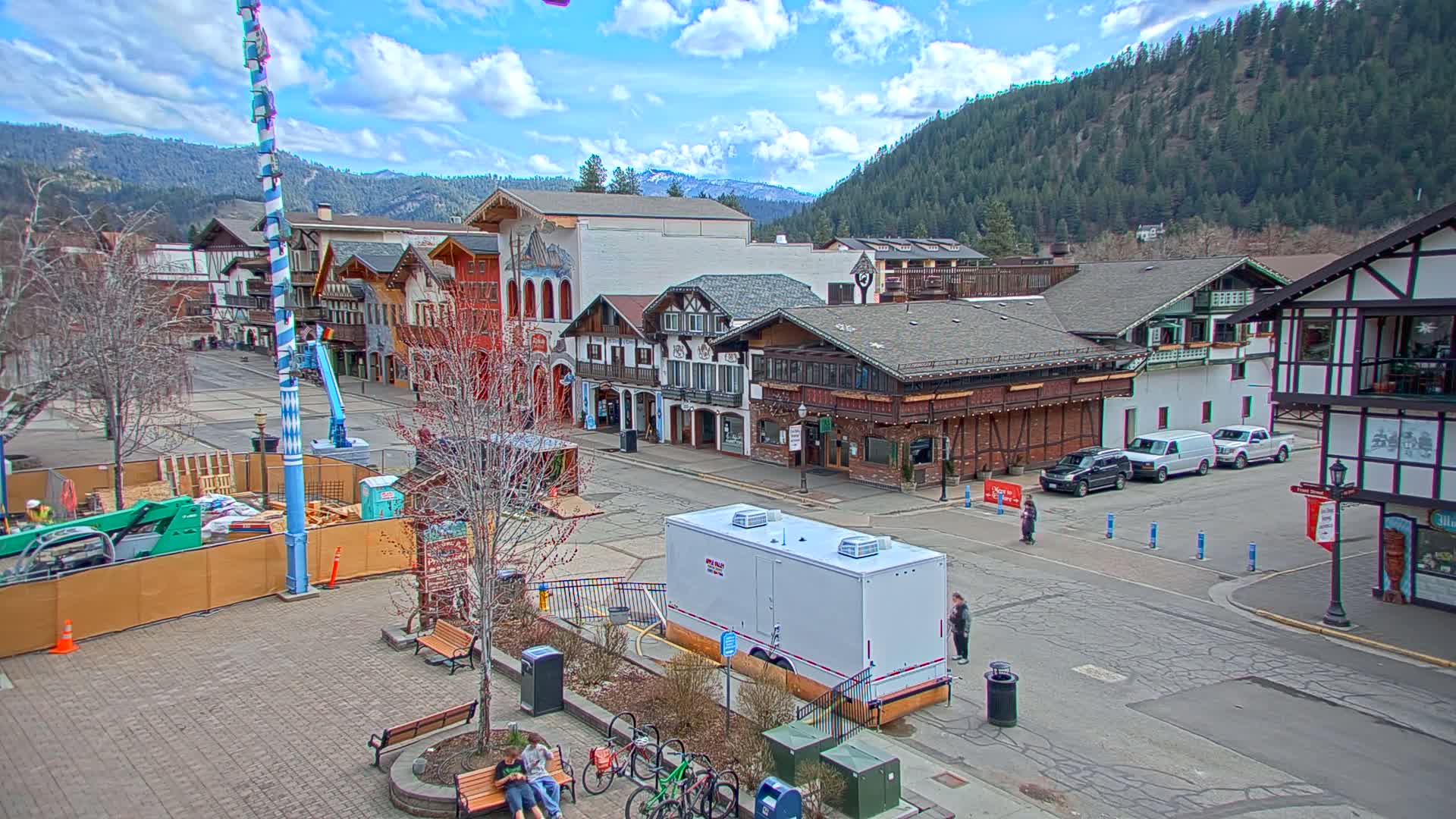 A town square with Bavarian-style buildings under a partly cloudy sky, construction underway in the square, and several vehicles parked nearby.