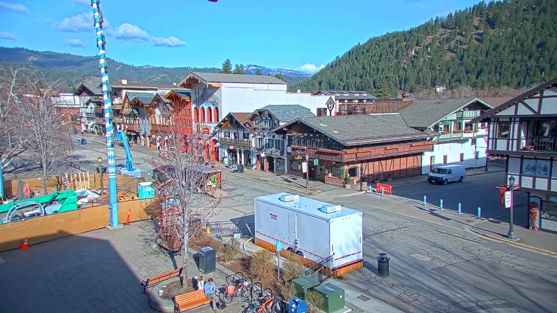 A sunny day reveals a town square with several Bavarian-style buildings, a construction area, and a few people sitting on a bench near bicycles; mountains are visible in the background.