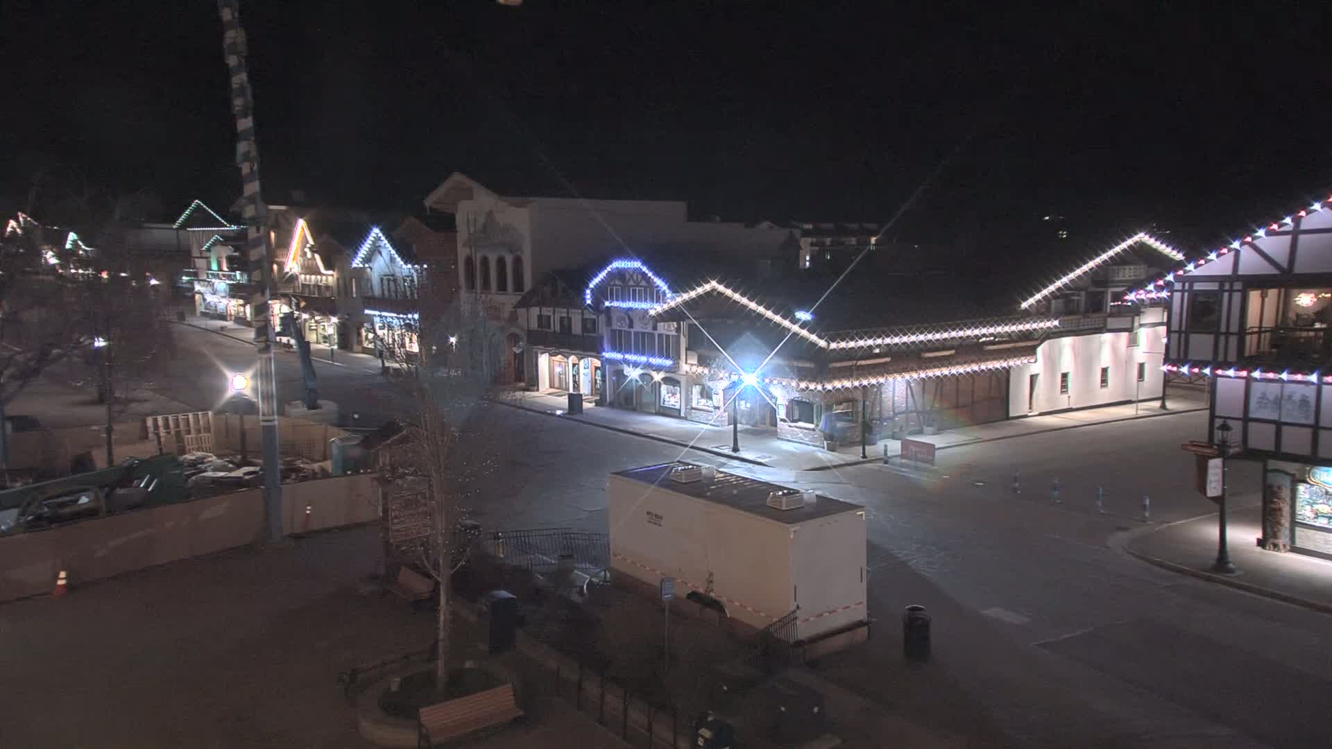 A mostly empty street at night lined with festively-lit buildings, showing some construction activity in the foreground.