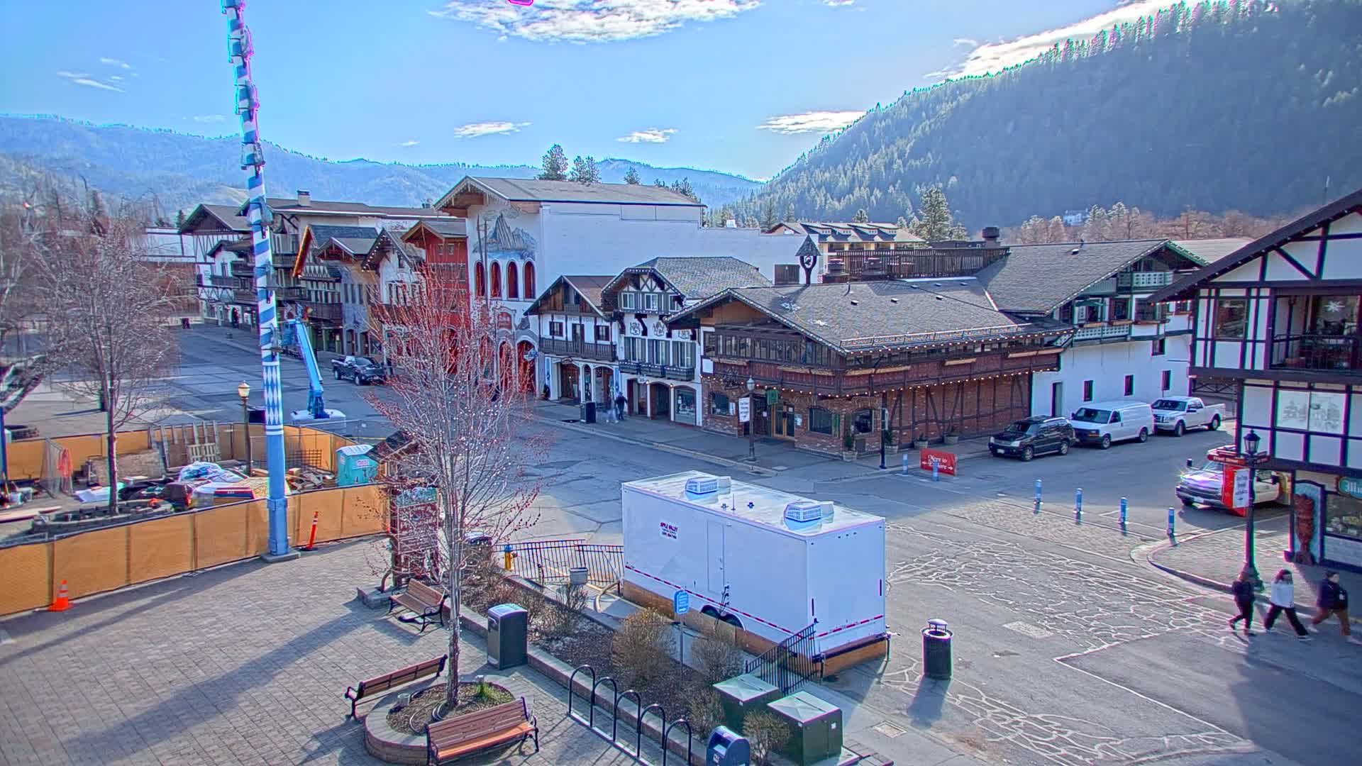 A town square with several buildings in a Bavarian architectural style sits under a sunny sky, with mountains in the background and construction underway in a section of the square.